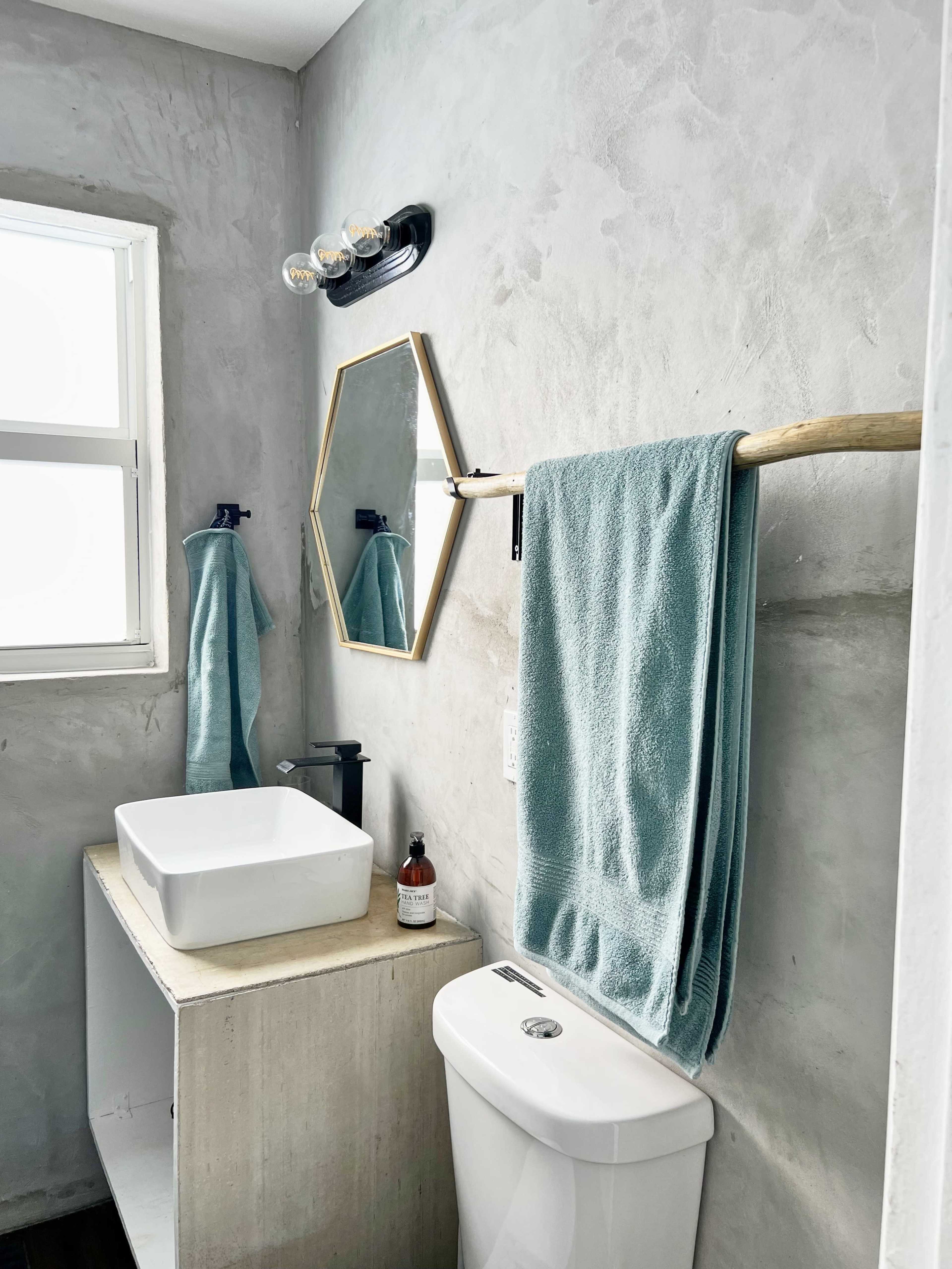 The image shows a modern bathroom featuring a concrete wall, a white square sink on a concrete vanity, a toilet, a towel rack with blue towels, and a hexagonal mirror above the sink.