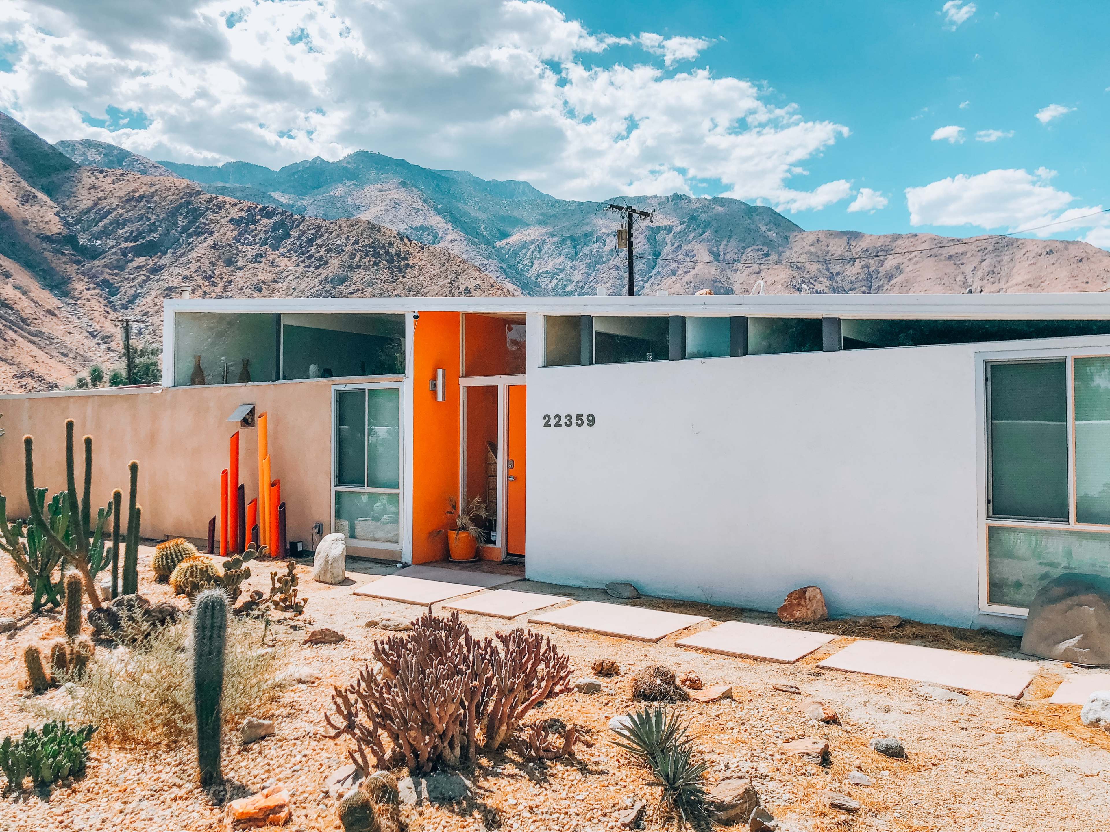 The image shows a modern house with an orange accent wall, set against a mountainous desert landscape with cacti and rocky terrain.