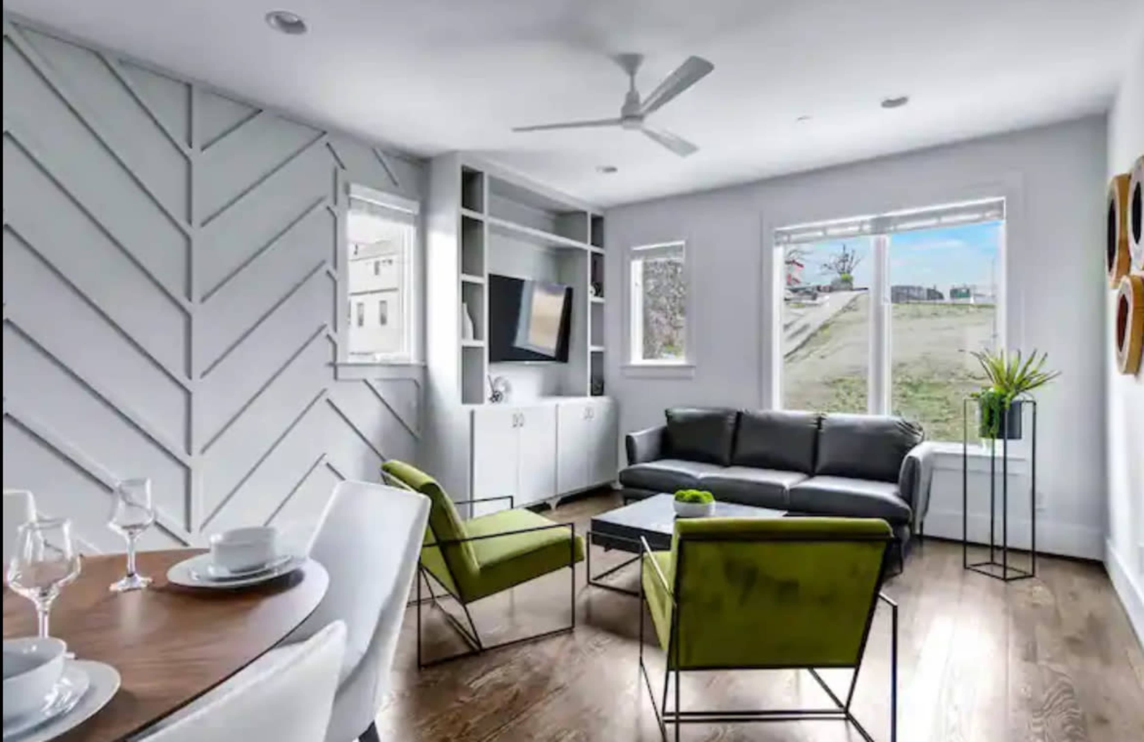 A modern living room with a black sofa, two green armchairs, a dining table set for two, and large windows that overlook a grassy area.
