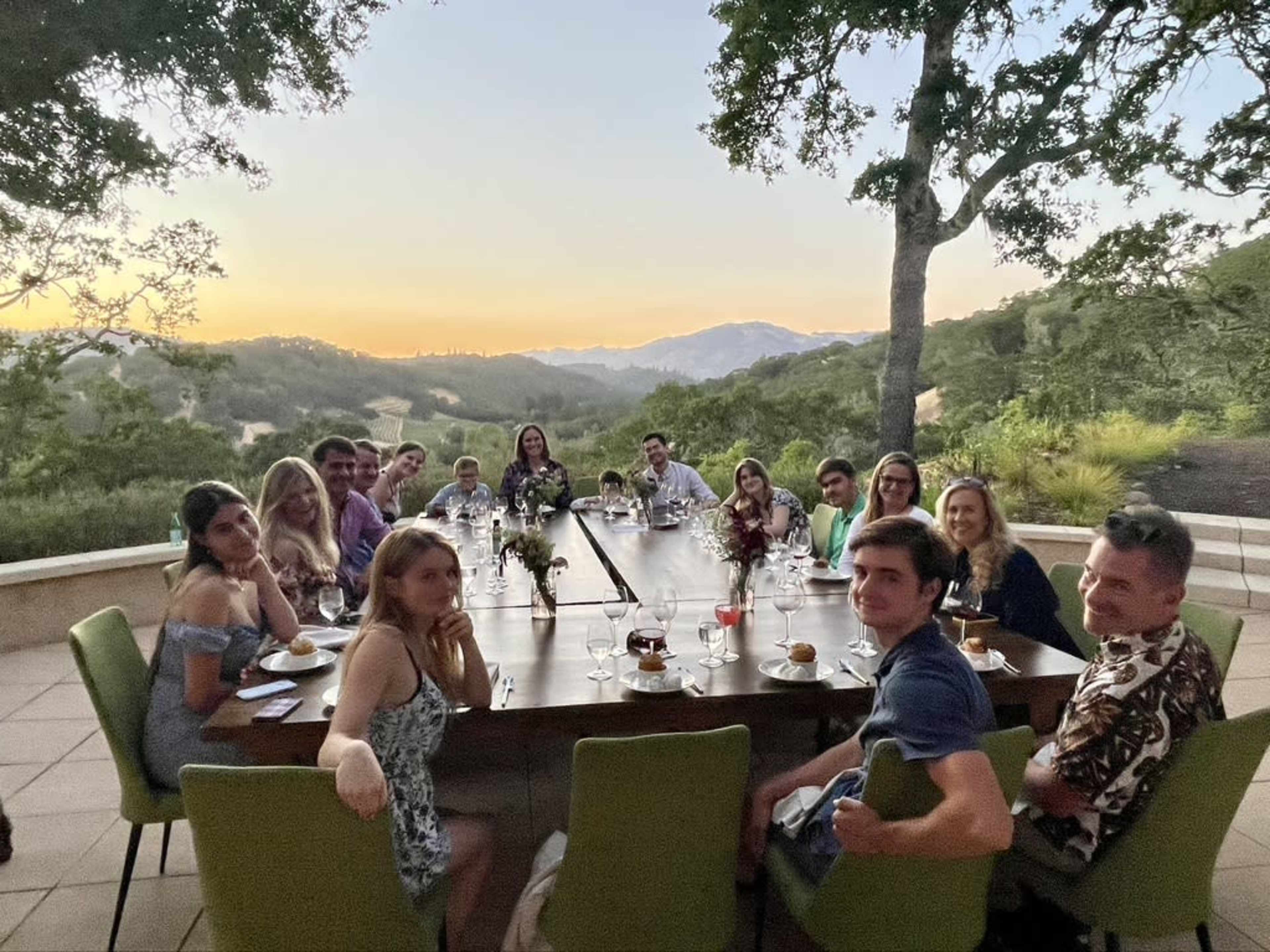 A group of people is seated around a long table outdoors, with a scenic view of hills and a sunset in the background.