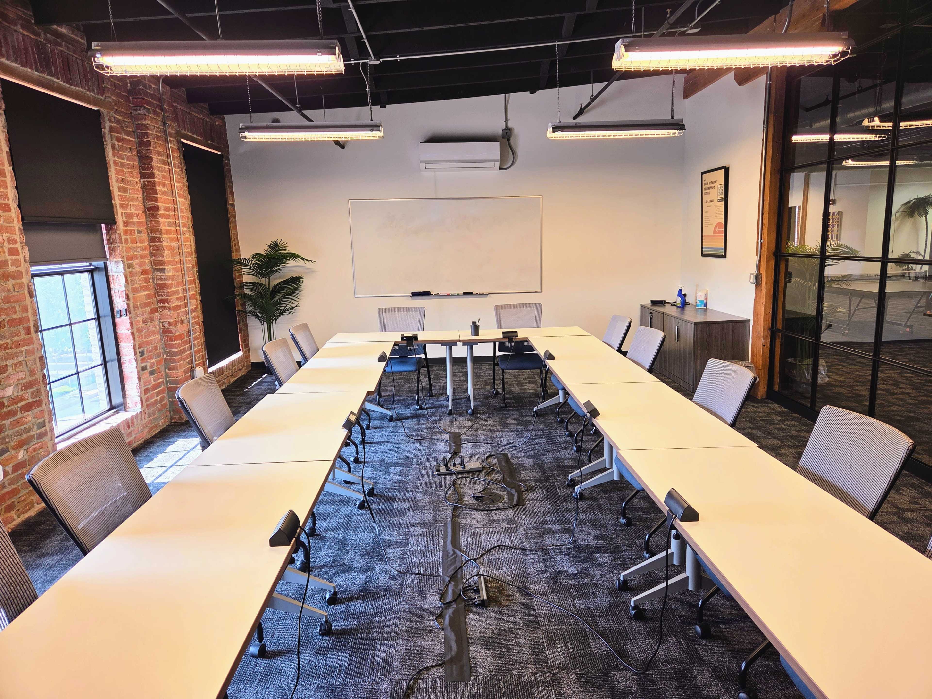 A conference room is arranged with a large oval table and multiple chairs, illuminated by overhead lights, with windows and a whiteboard visible on the walls.