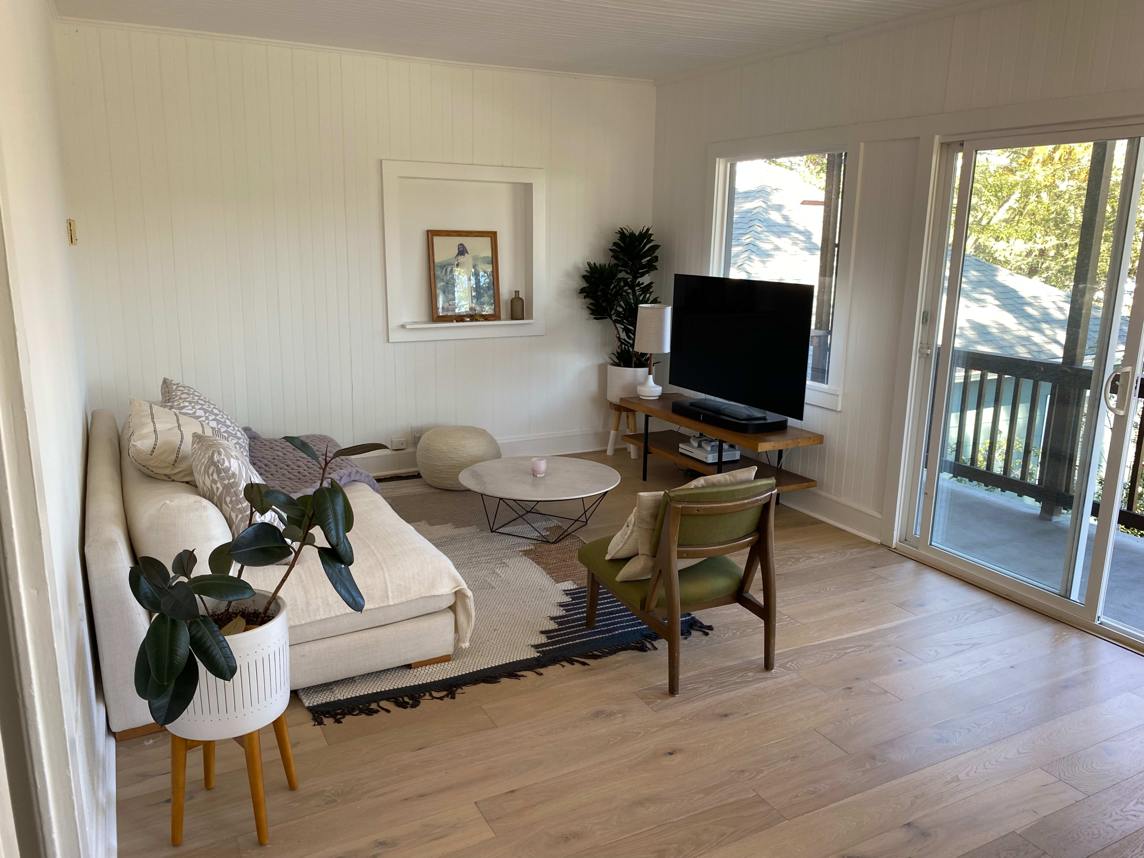 A bright living room with a white sofa, a round coffee table, a television on a stand, and large windows overlooking a balcony.