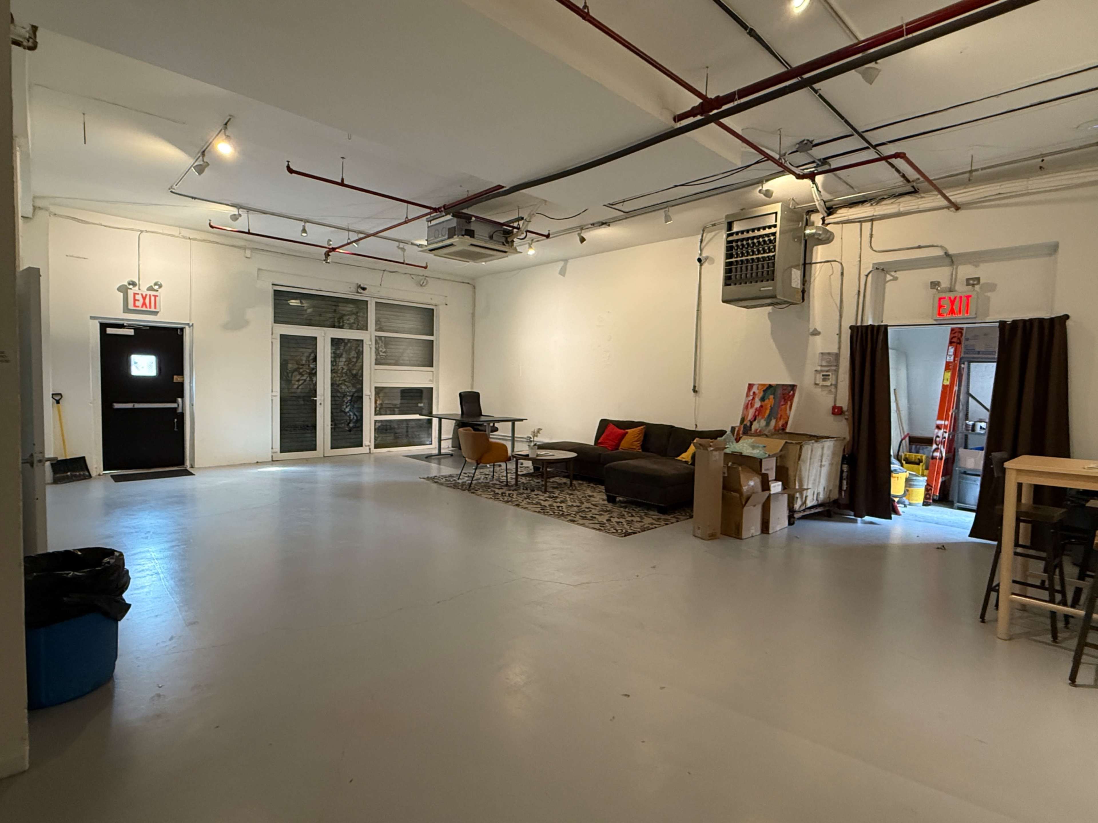 The image shows a spacious, empty room with concrete flooring, large windows, and various furnishing elements, including a sofa, a table, and storage boxes.