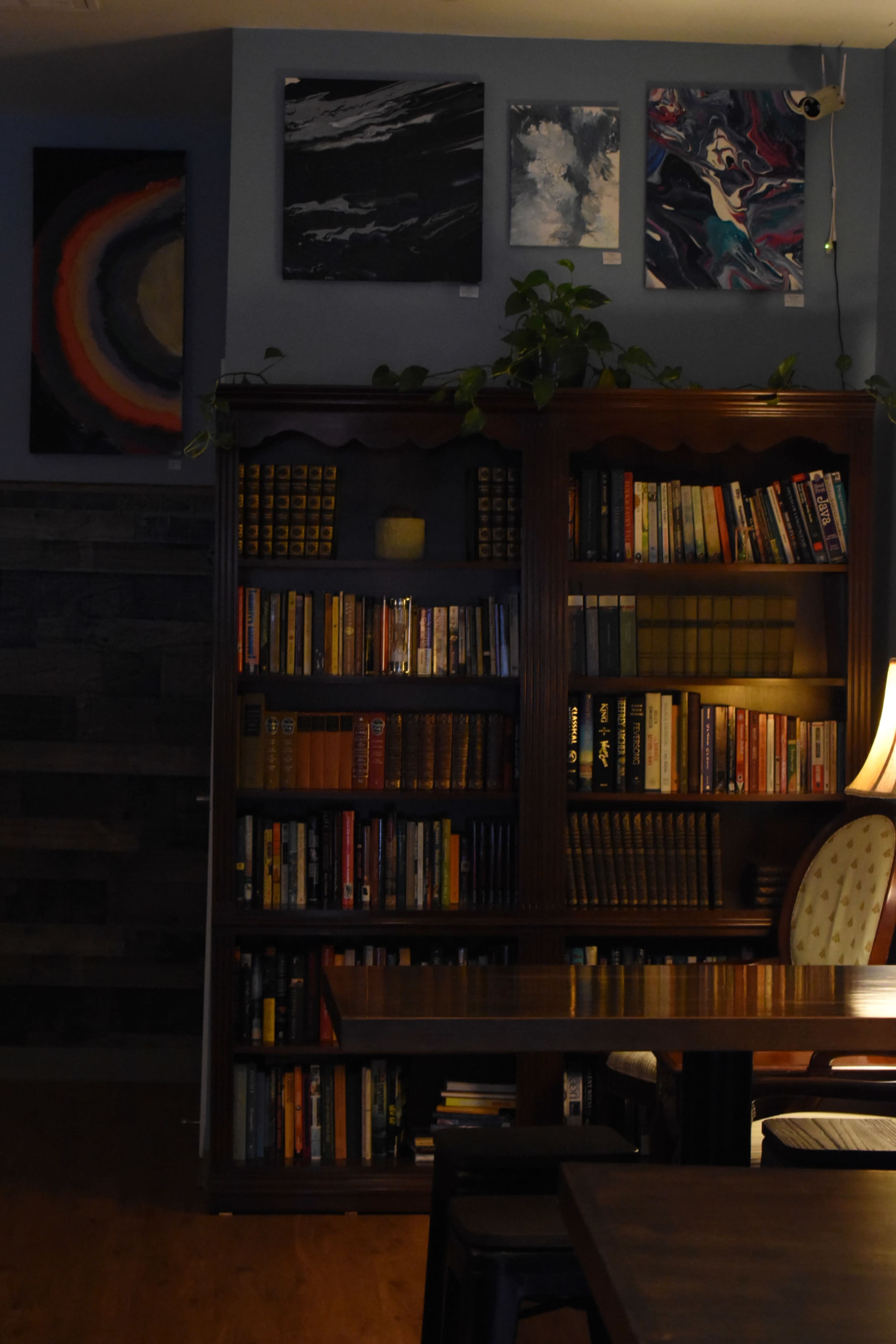 A dimly lit room with a bookshelf filled with various books, alongside paintings on the wall and a lamp illuminating a nearby wooden table.