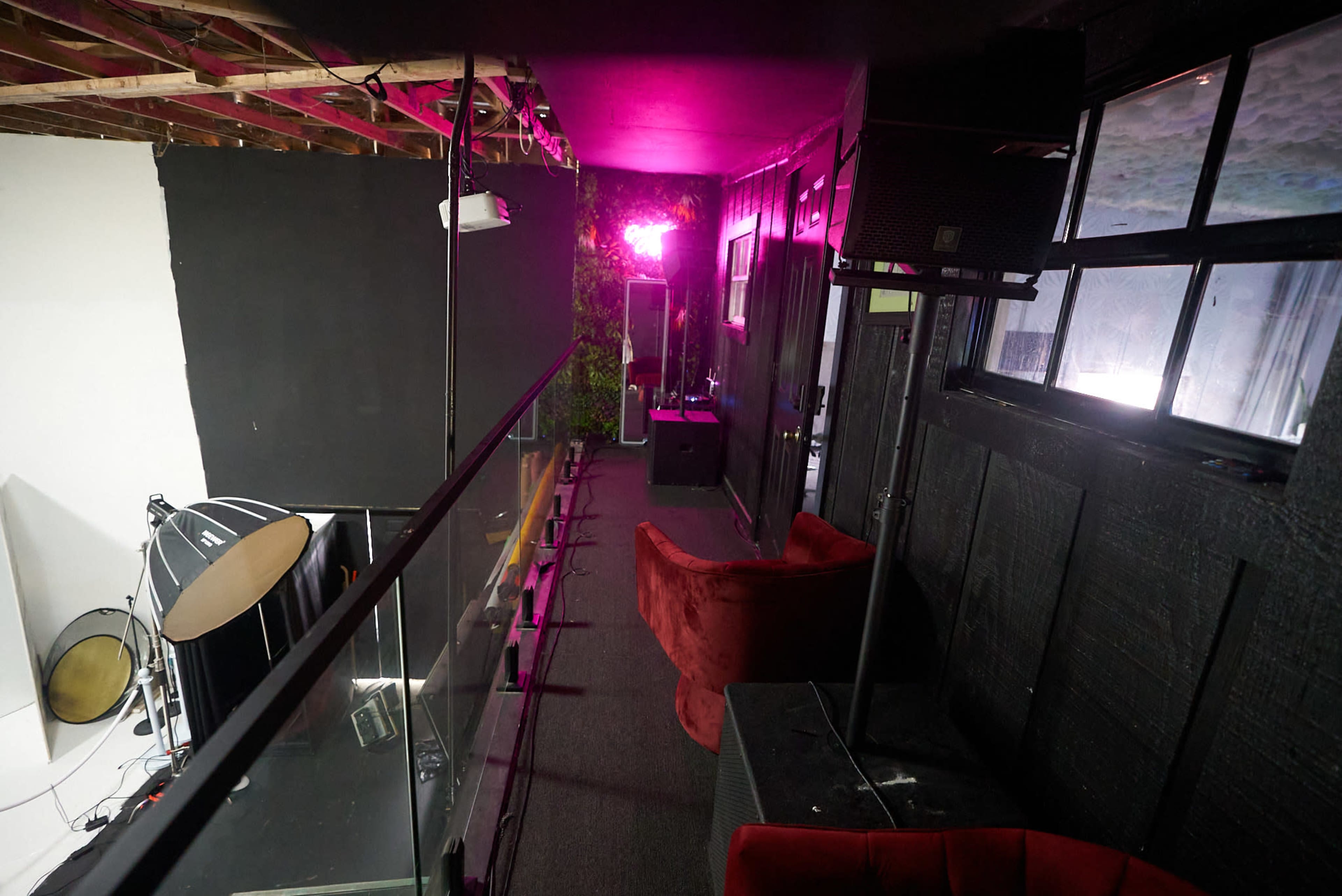 The image shows a balcony area with red seating, overlooking a room lit by pink lights, and featuring equipment such as musical instruments and speakers.