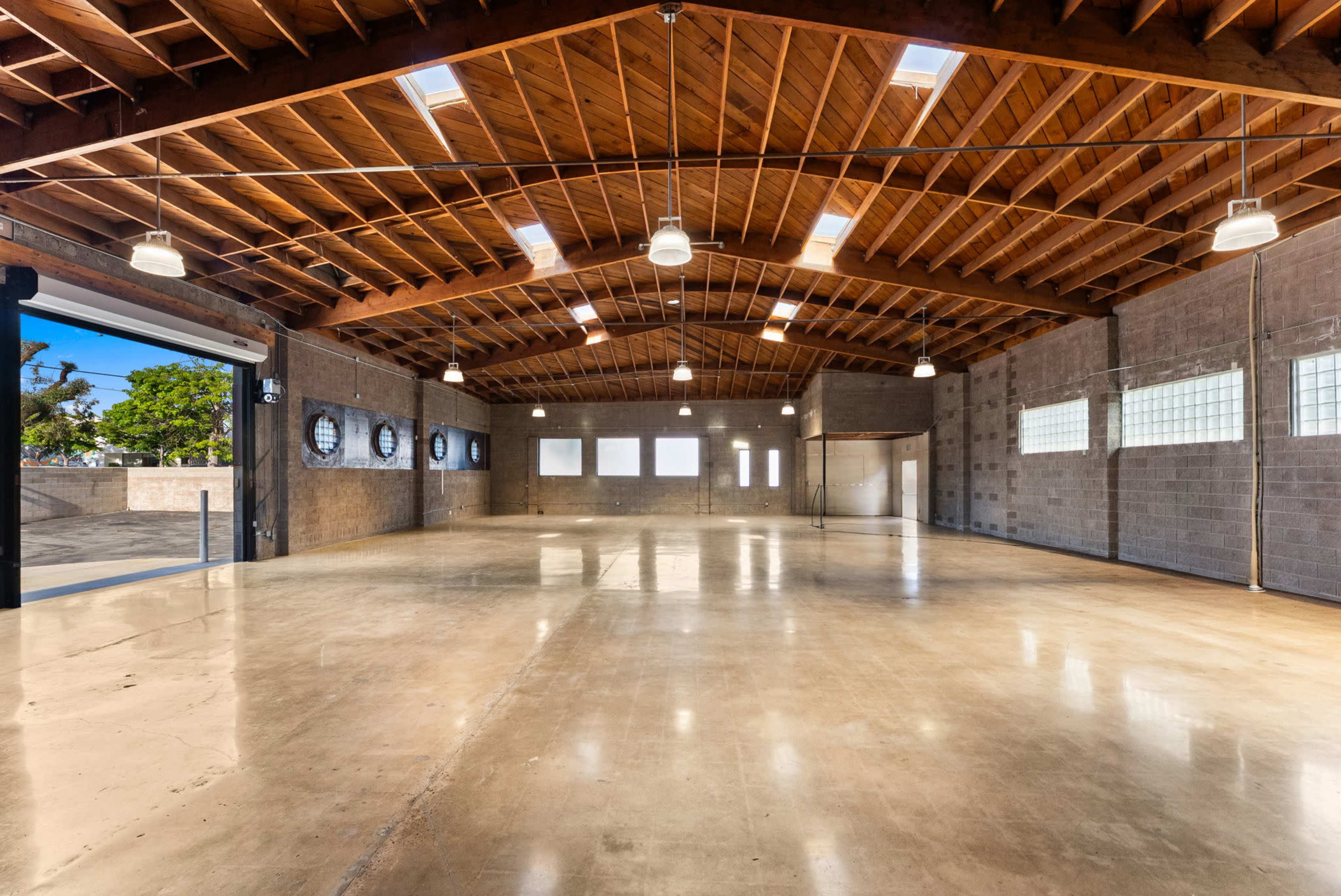 The image shows a spacious, empty indoor area with high wooden beams, concrete walls, and a polished floor.