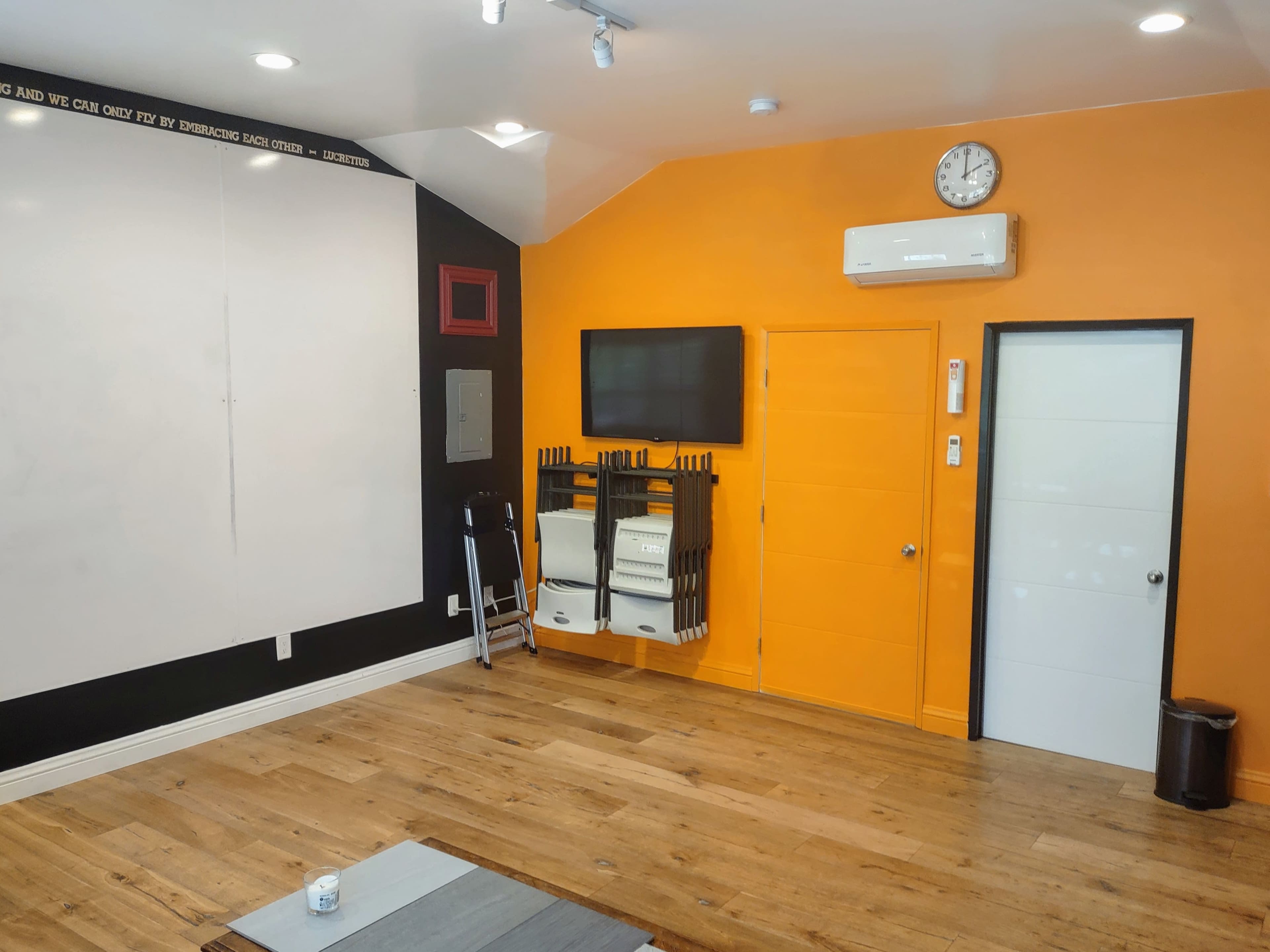 A brightly colored room with an orange accent wall, a whiteboard, wooden flooring, and a mounted TV along with a door leading to another space.