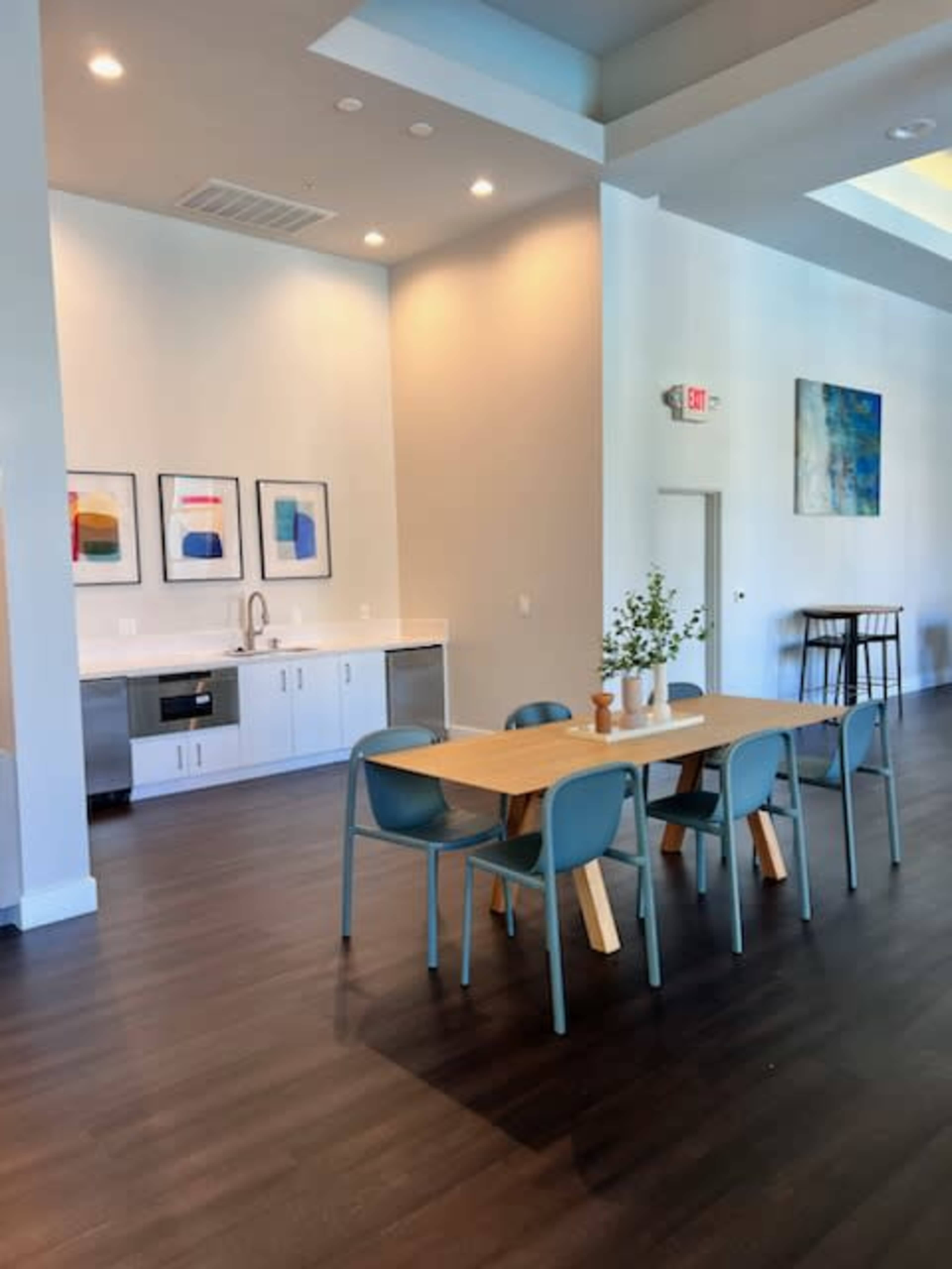 A modern dining area features a wooden table surrounded by blue chairs, with a kitchenette and framed artwork on the walls.