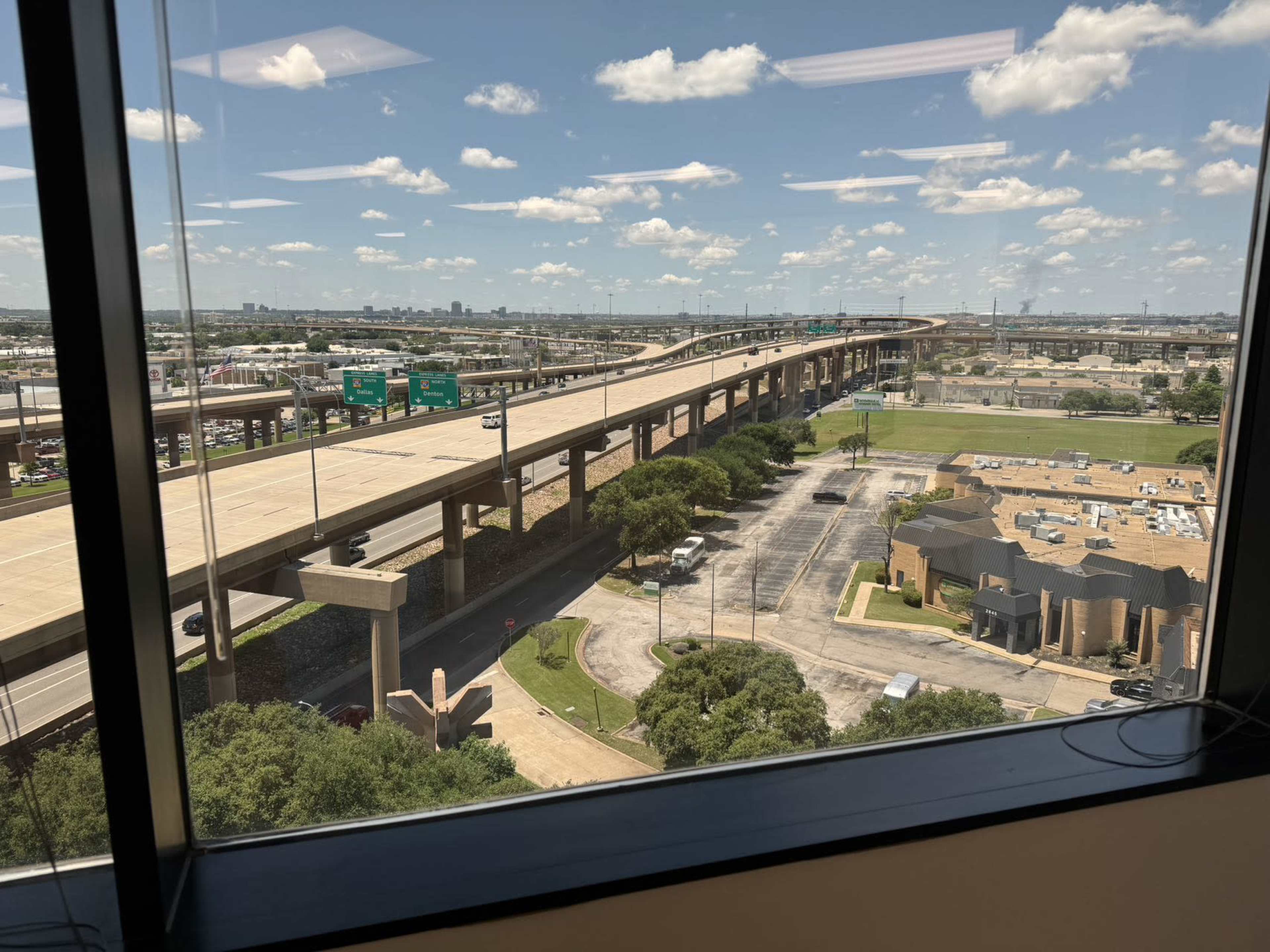 A highway overpass viewed from a window, with a bustling urban landscape in the background, including buildings and roads.