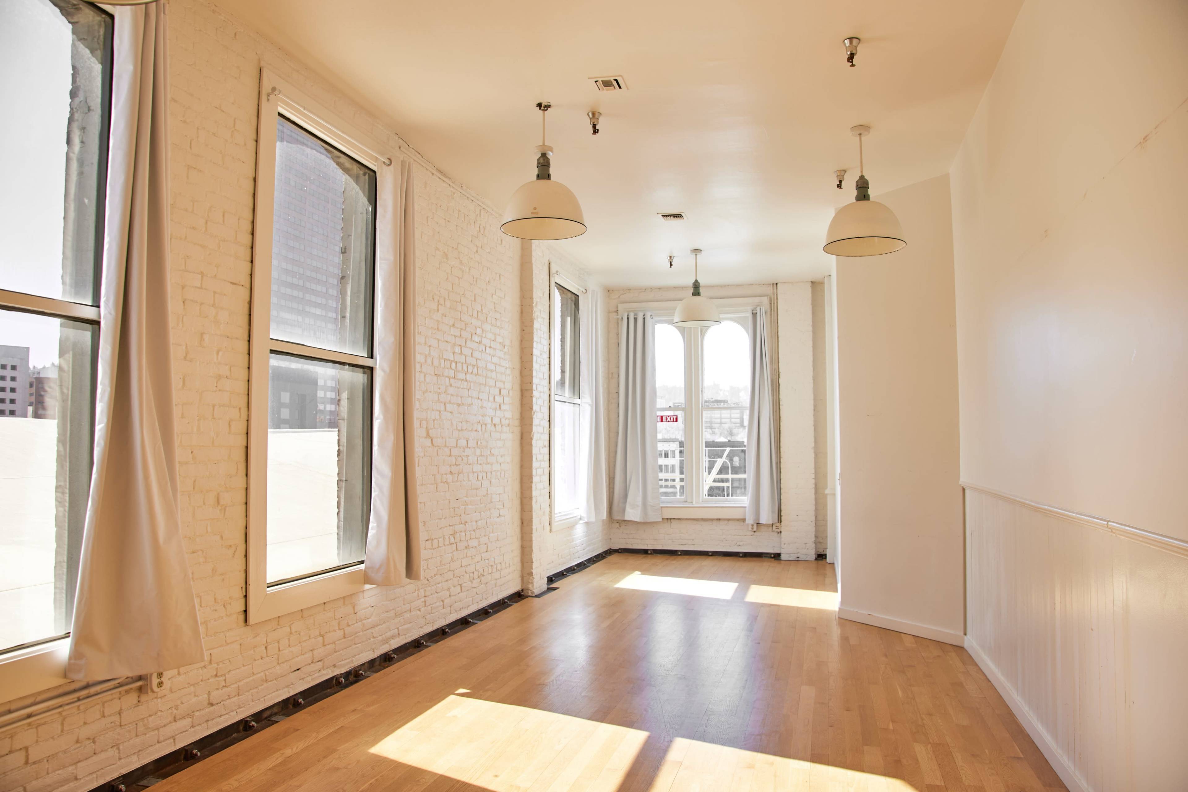 A spacious, sunlit room with large windows and wooden flooring, featuring exposed brick walls and industrial-style pendant lights.