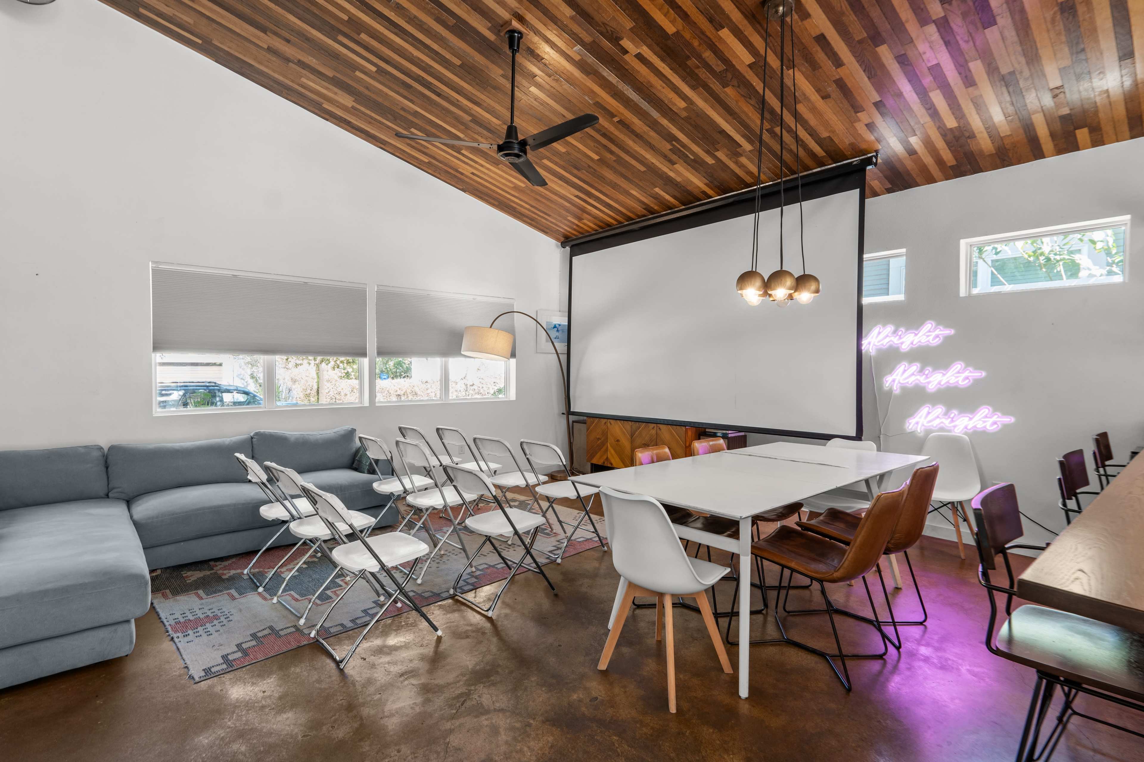 The image shows a modern meeting space with a mix of seating arrangements, including folding chairs and a long table, under a wooden ceiling.