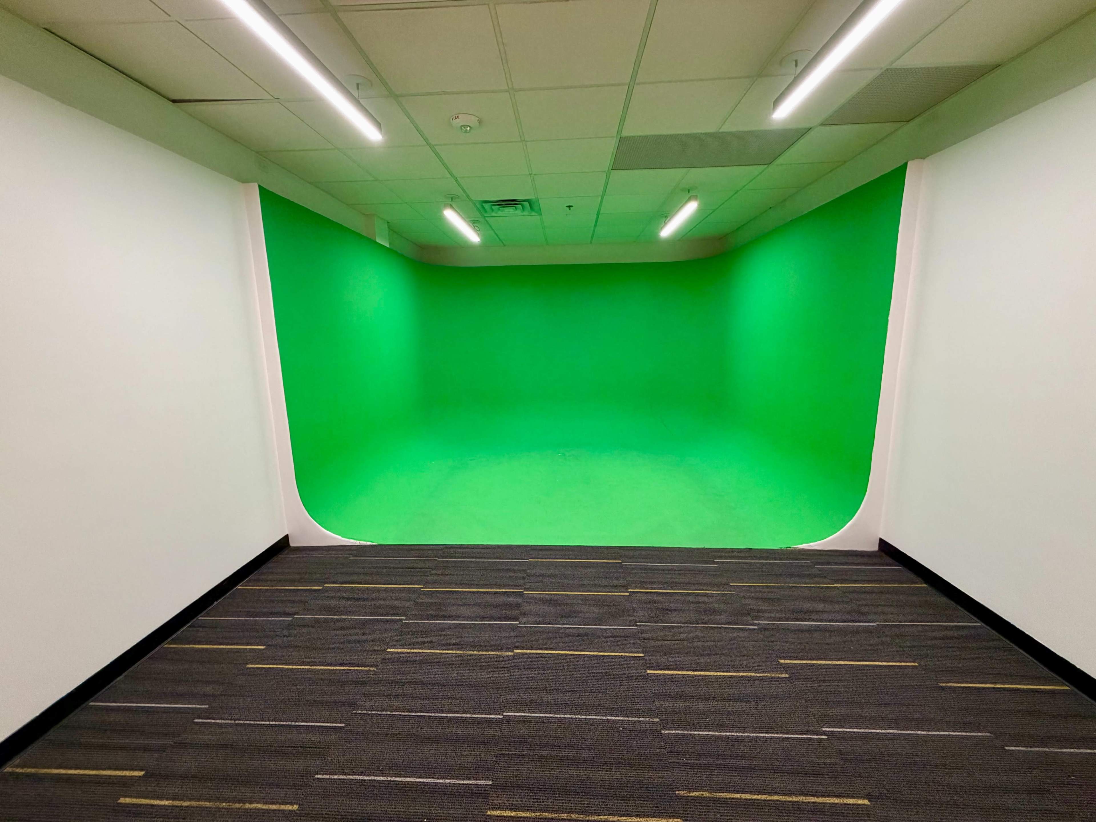 The image shows a room with a green screen backdrop, surrounded by white walls and a gray carpeted floor.