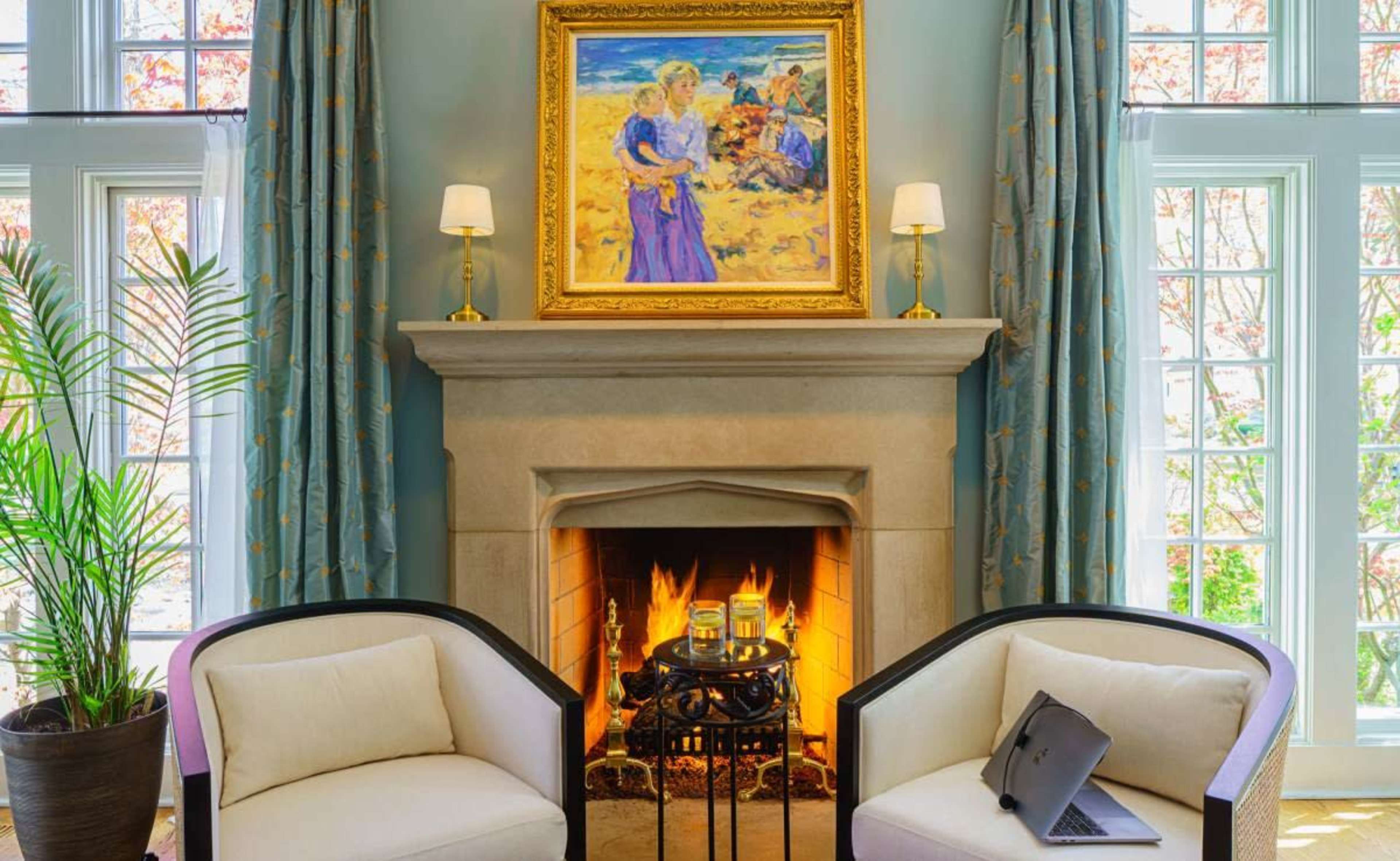A cozy living room features two armchairs, a decorative painting above a stone fireplace, and a warm fire burning inside.