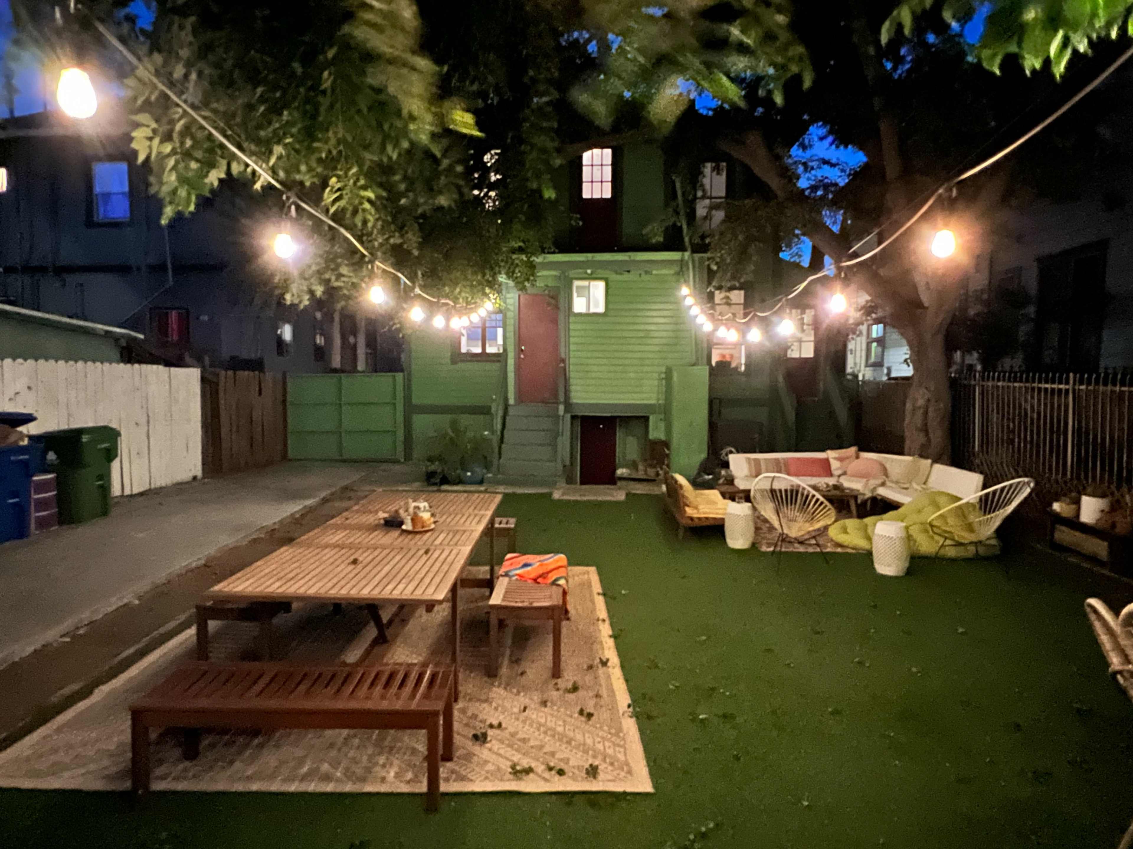 A green two-story building in a backyard with a wooden dining table, lounge chairs, and string lights illuminating the evening scene.