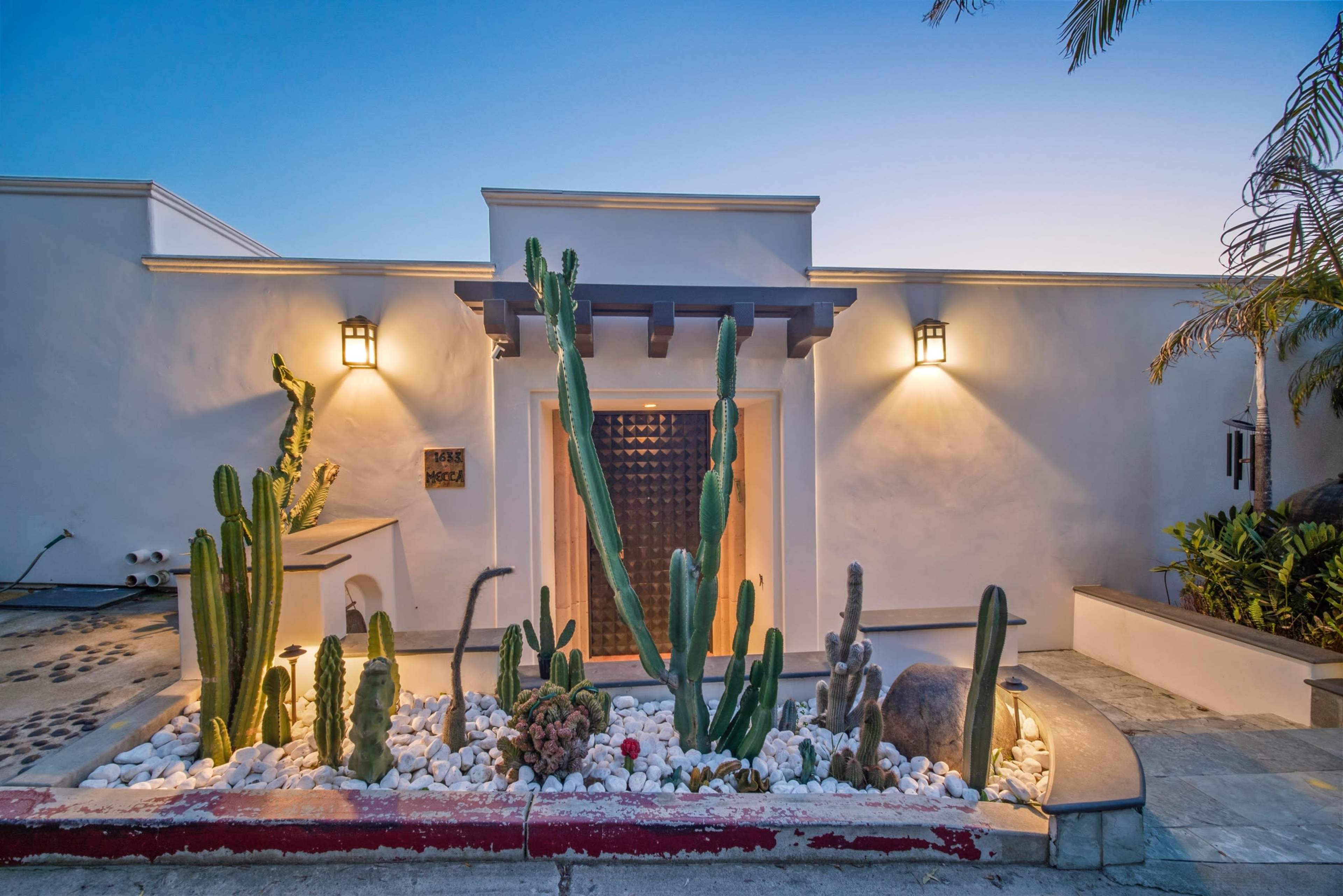 A modern entrance features a wooden door surrounded by a garden of cacti and white stones, illuminated by wall sconces.