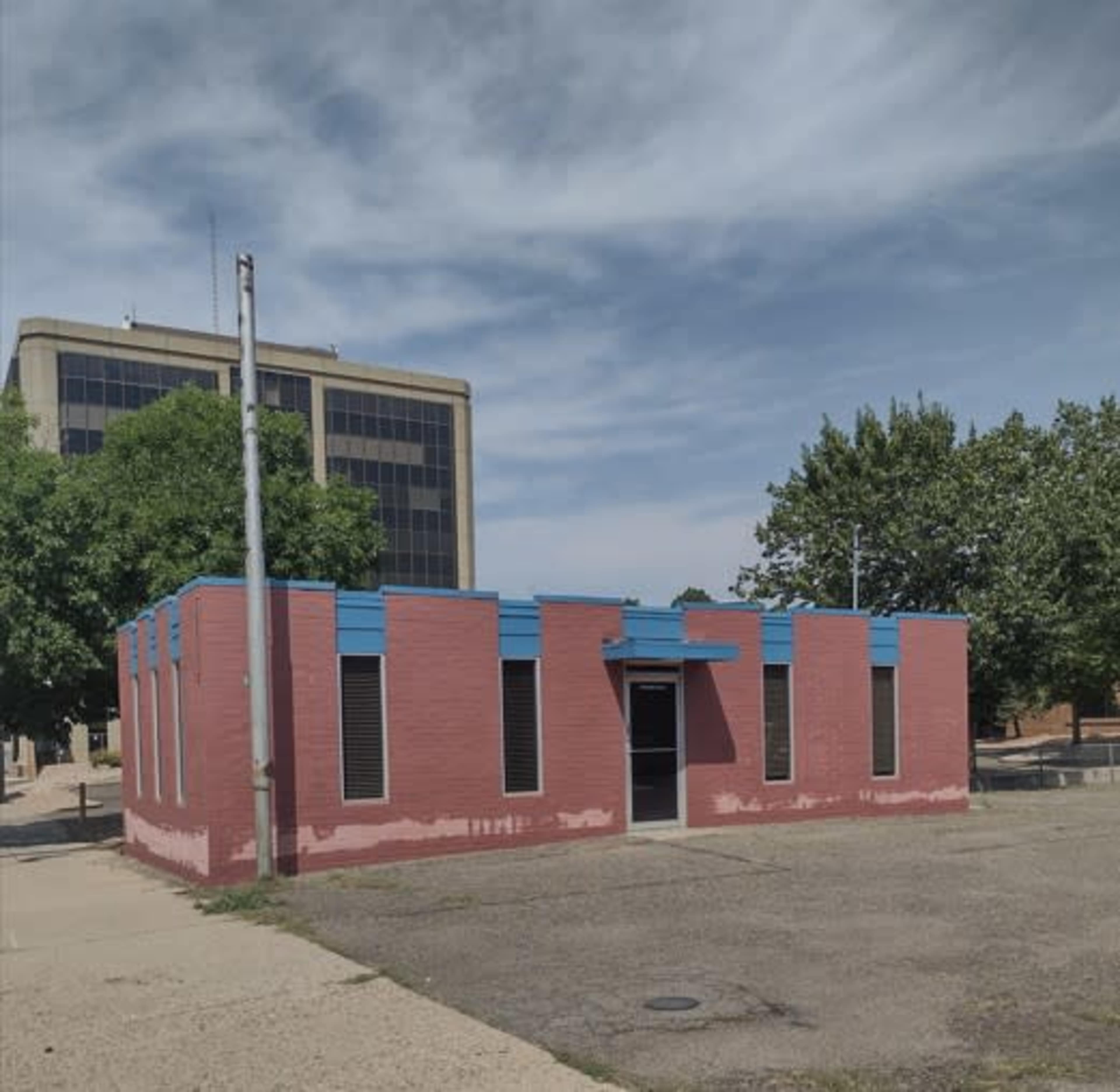 A one-story, pink building with blue accents is situated next to a taller building and surrounded by trees and an empty parking lot.