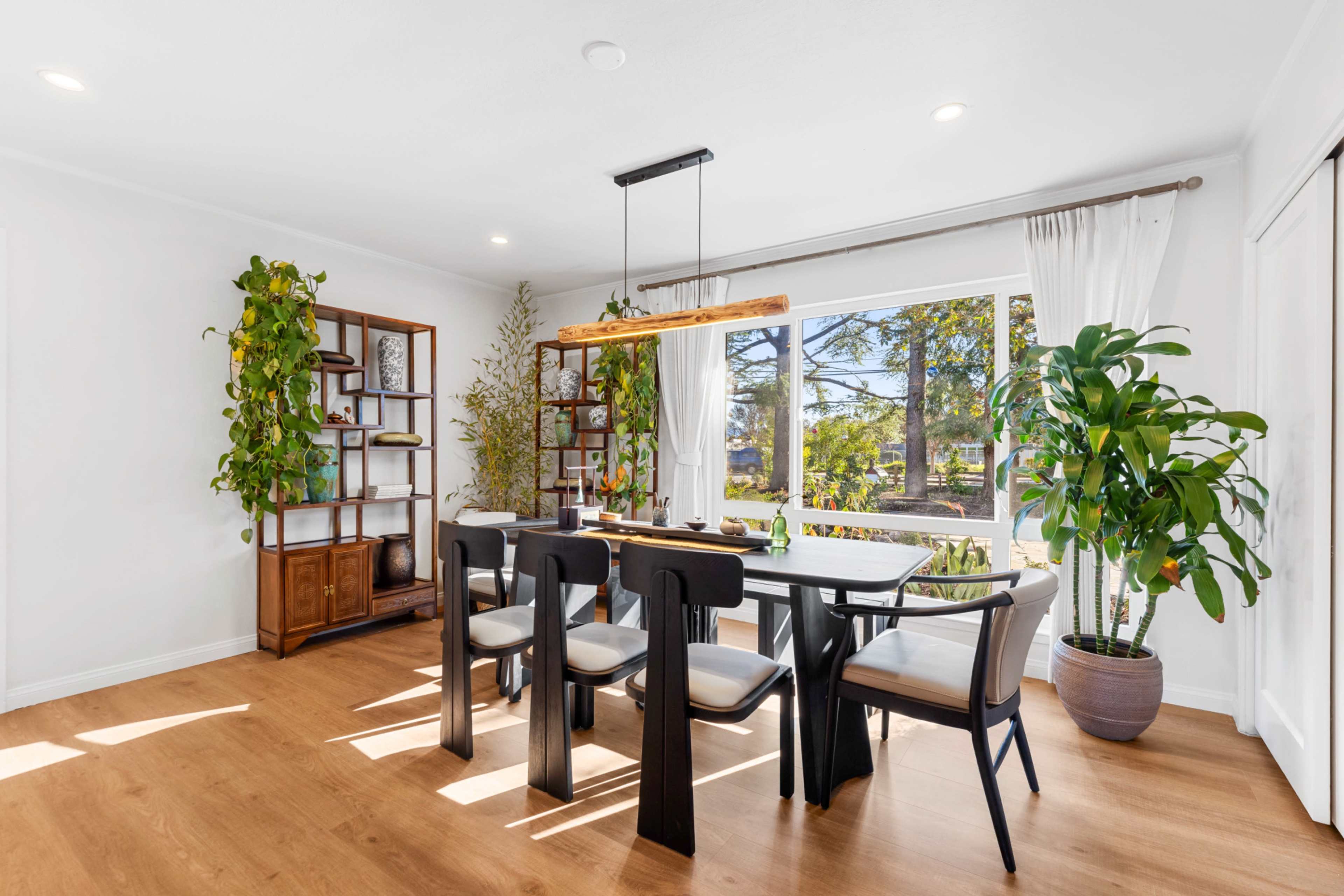 The image shows a modern dining area with a wooden table, six chairs, and large windows allowing natural light, surrounded by potted plants and decorative shelving.