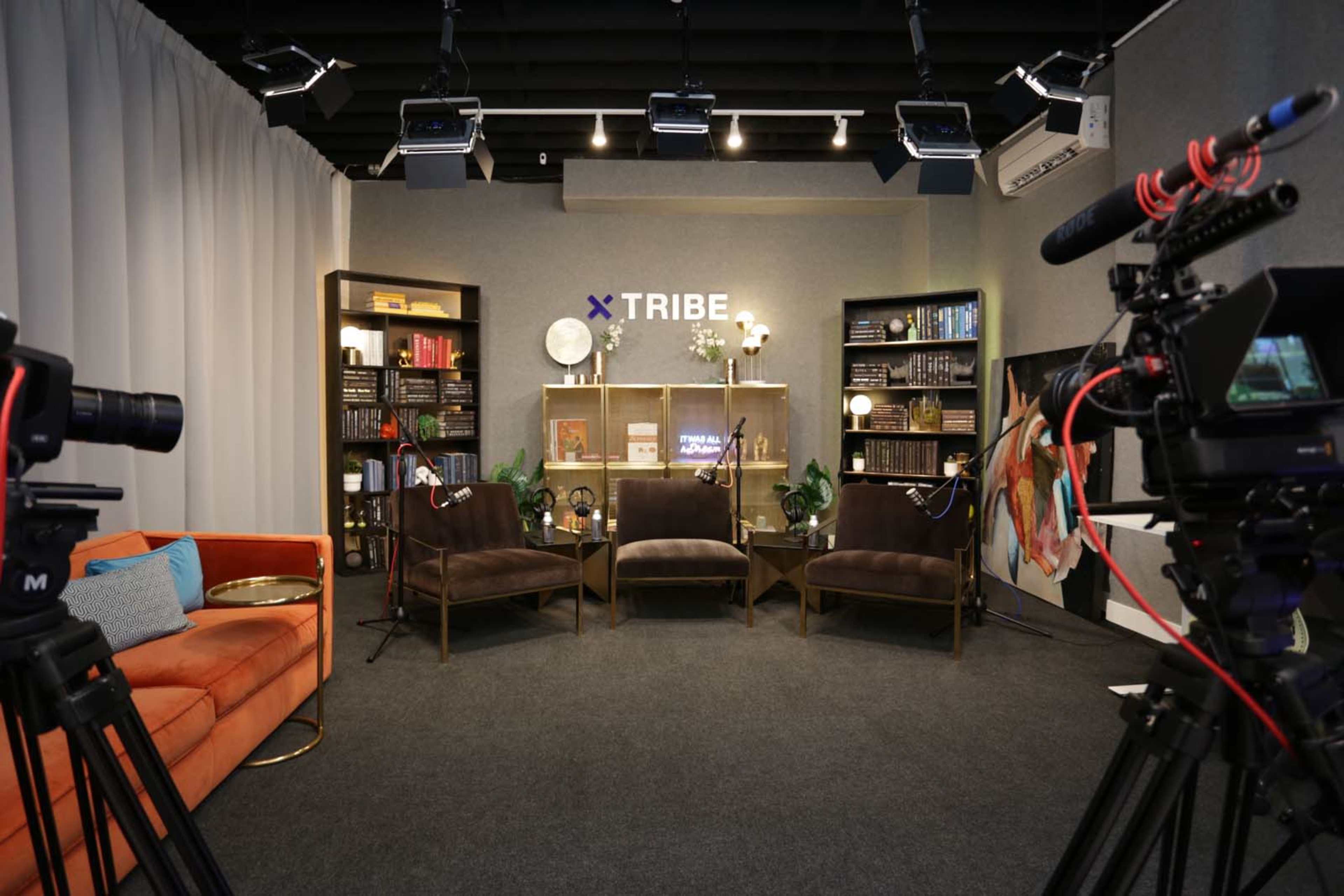 A broadcast studio set with two brown chairs and an orange couch, surrounded by cameras and decorative bookshelves.