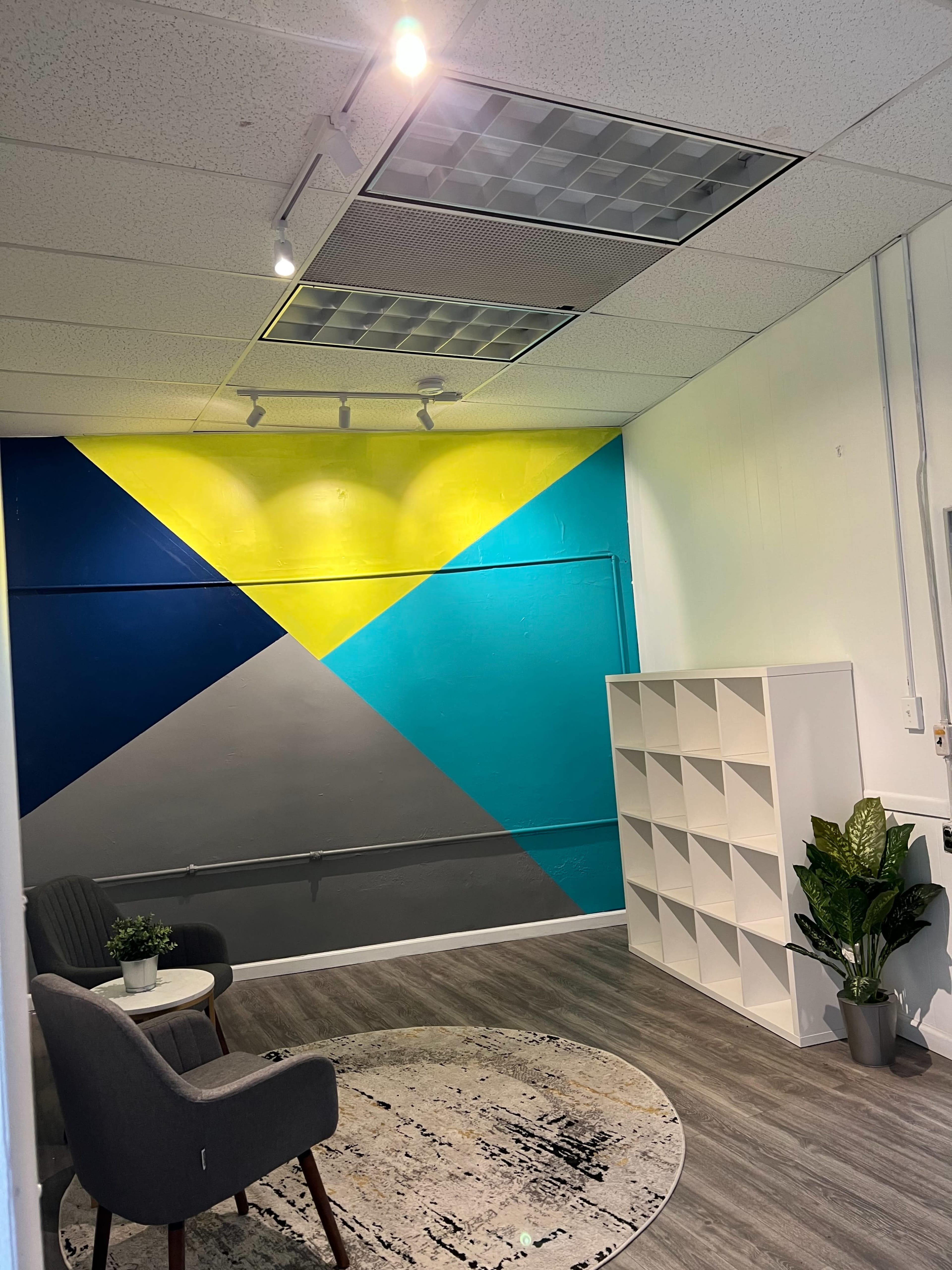 A modern room with a geometric mural on the wall, a white shelving unit, a small plant, and two chairs arranged in a seating area.