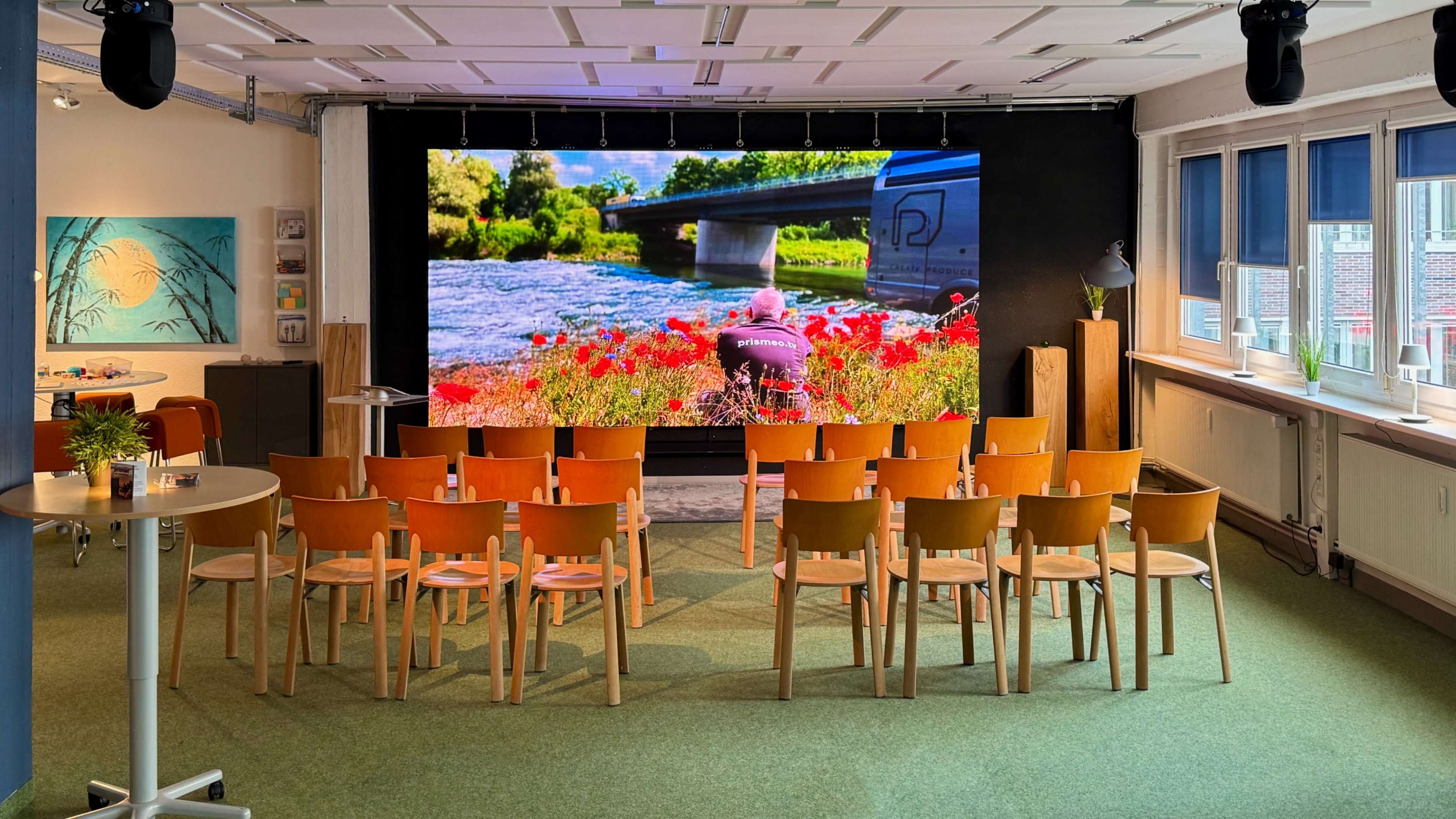 A row of chairs faces a large screen displaying a vibrant landscape scene with a river and flowers.