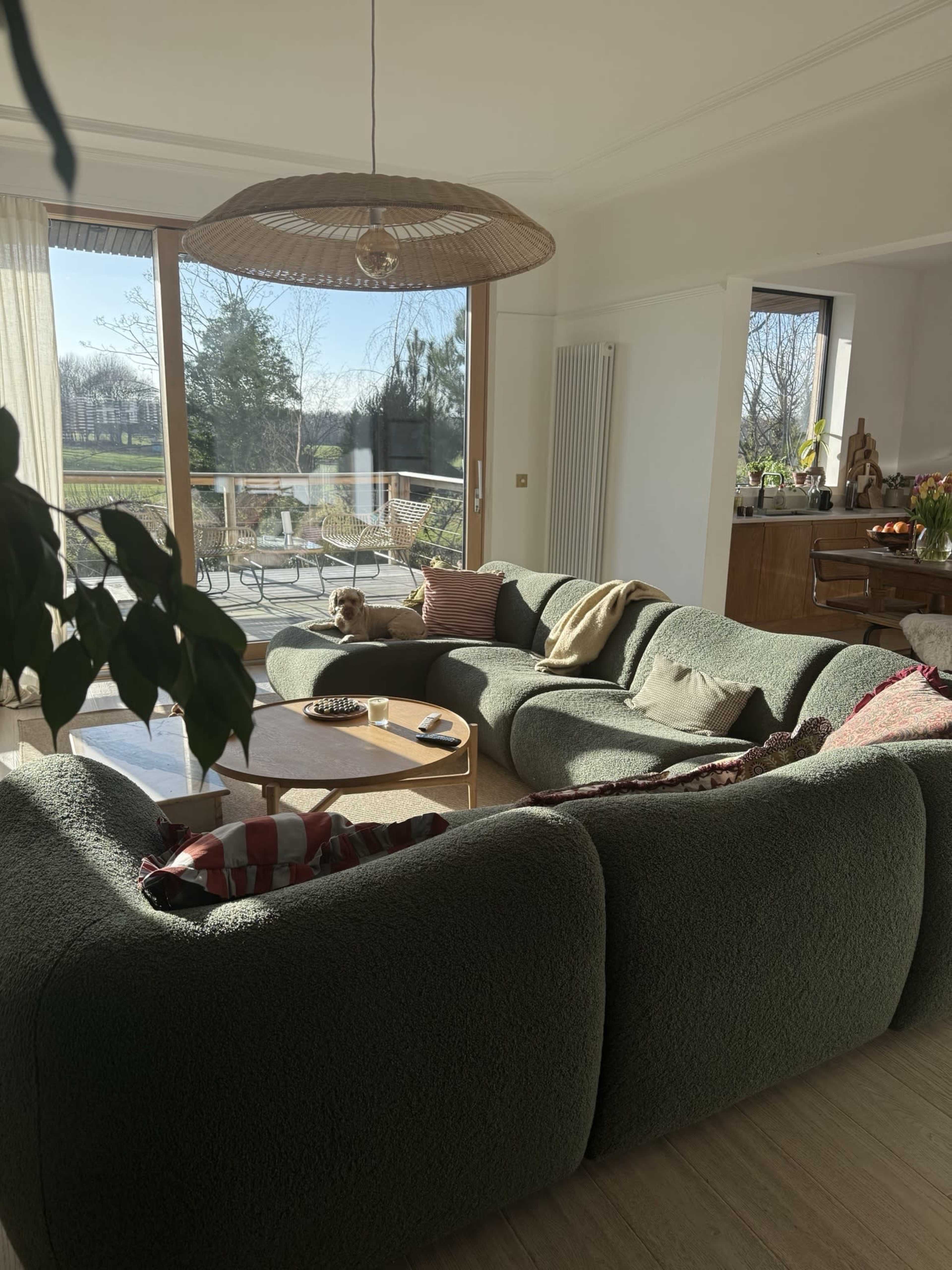 The image shows a cozy living room with a large, curved green sofa, a wooden coffee table, and a view of an outdoor patio through large windows.