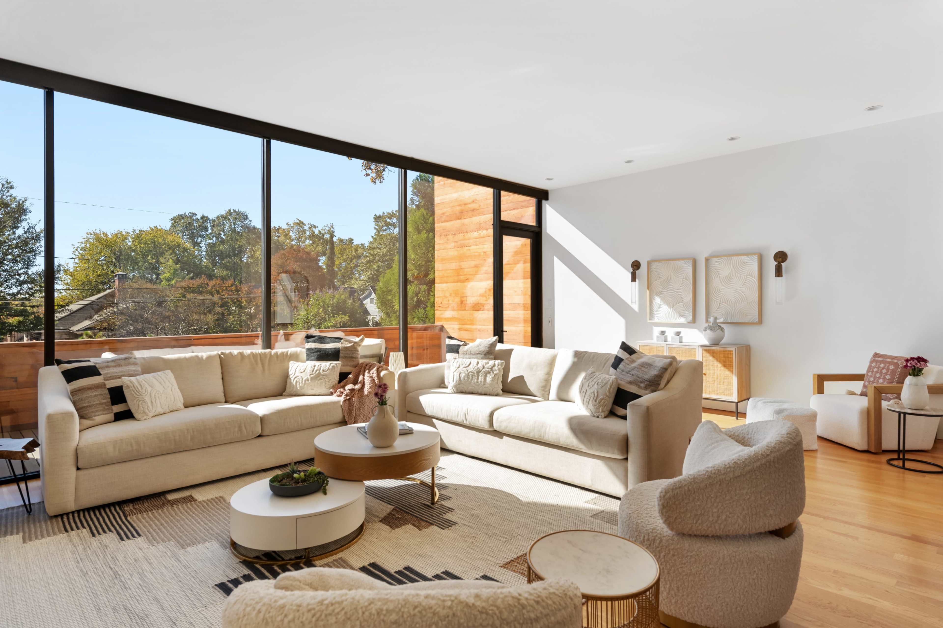 The living room features large windows, two beige sofas, a round coffee table, and a neutral color palette with wooden accents.
