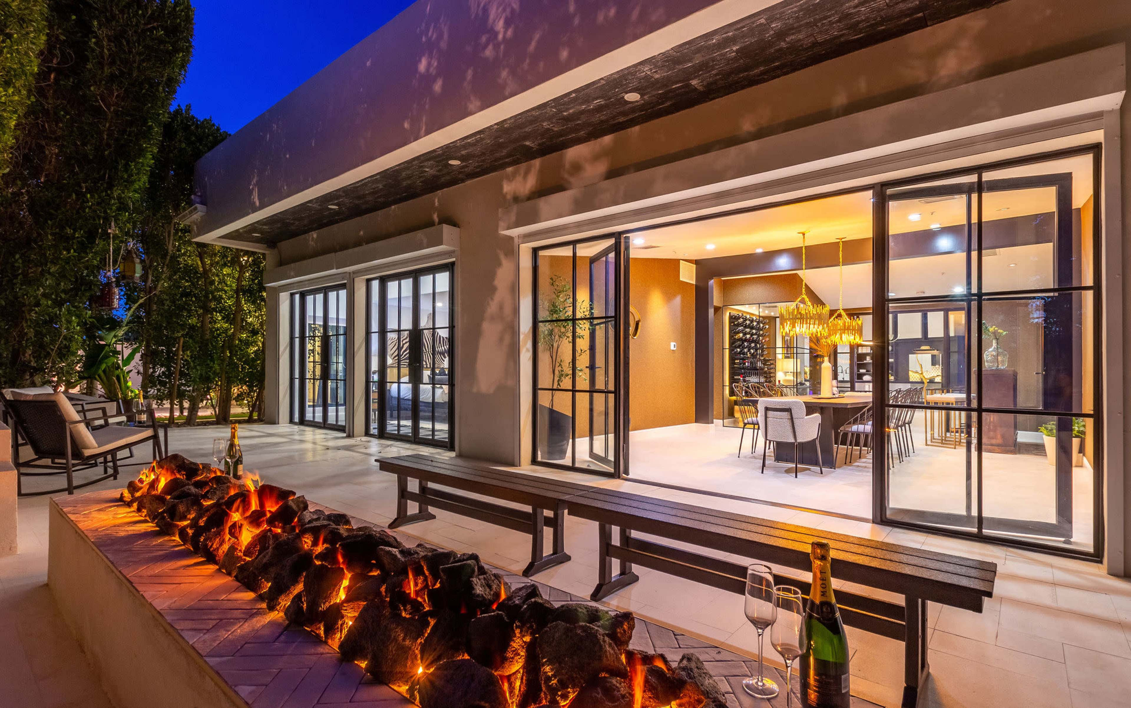 A modern outdoor living space with a fire feature, surrounded by greenery, leading to an interior dining area with large windows.