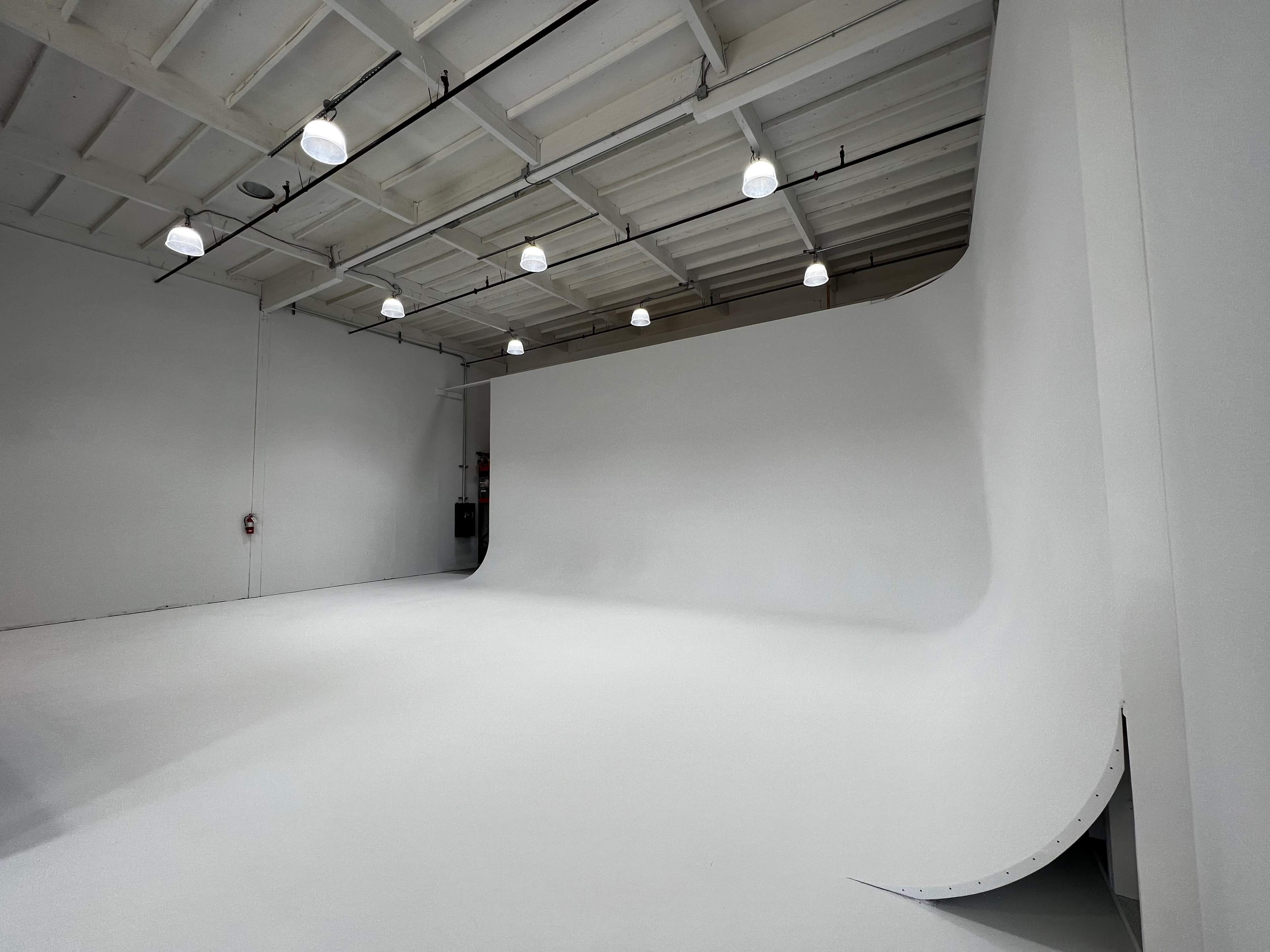 A spacious, empty photography studio with a white backdrop and overhead lights.