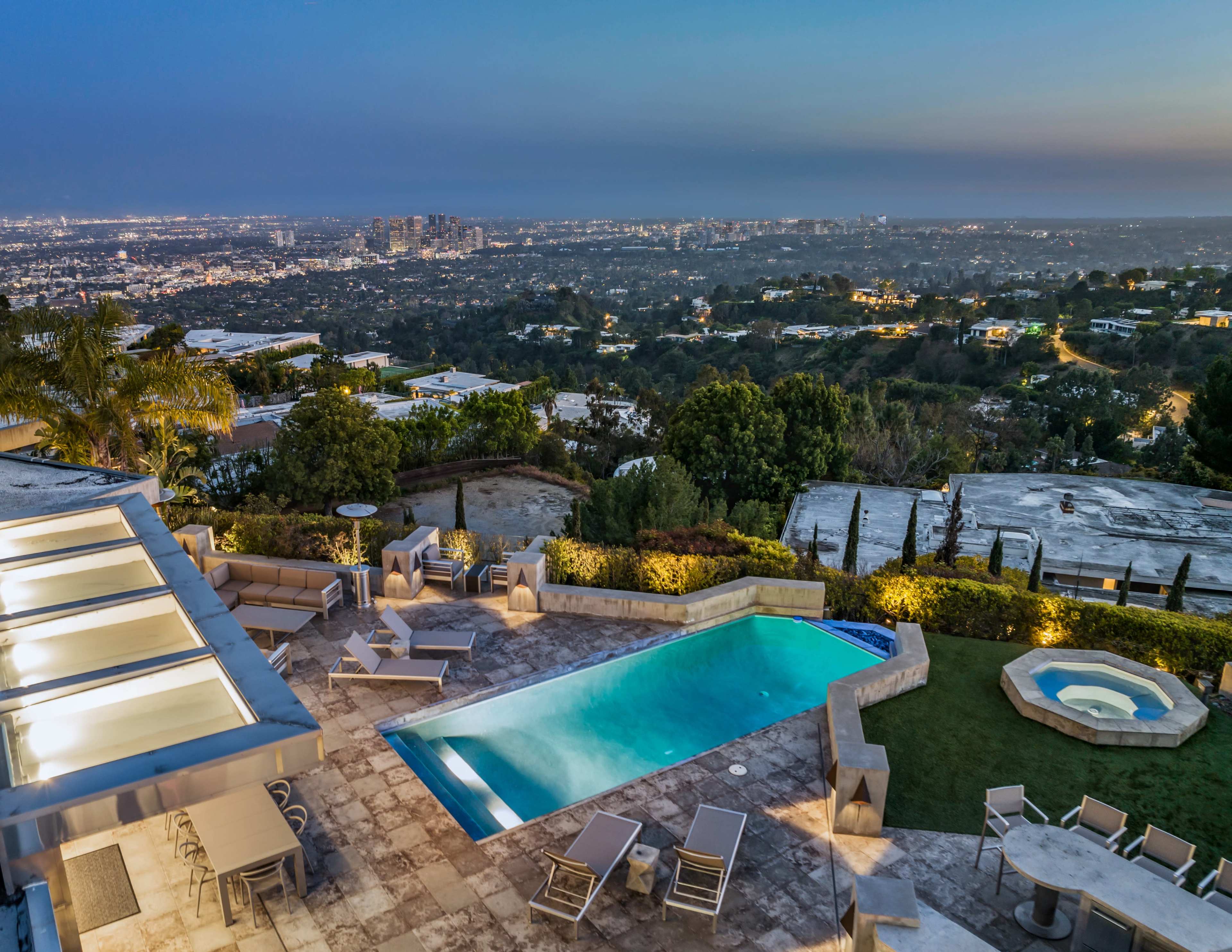 A hillside villa features a swimming pool, spa, and seating area with a panoramic view of the city skyline at dusk.