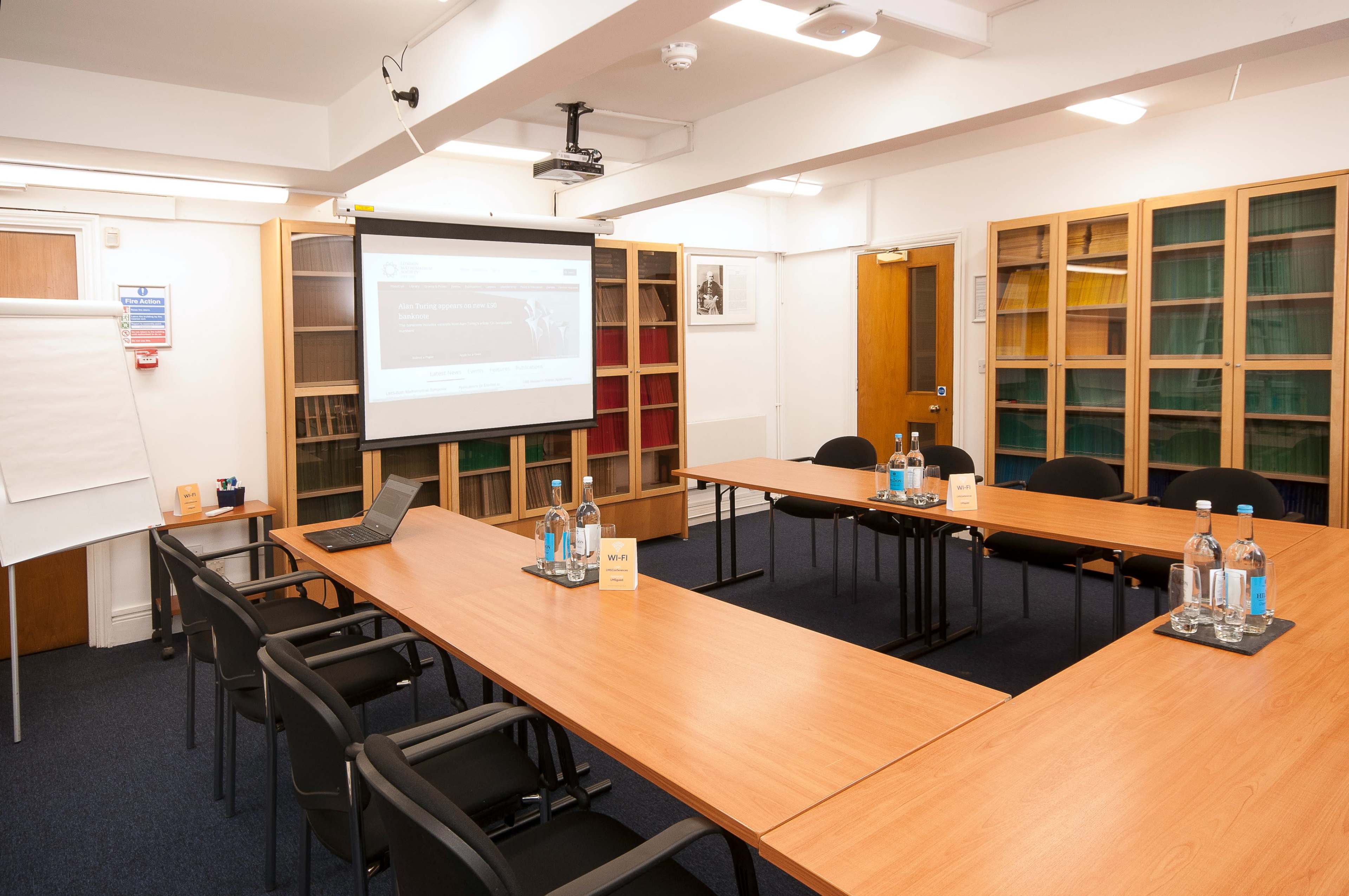 A conference room features a large rectangular table surrounded by black chairs, a projector screen at the front, and shelves filled with books in the background.