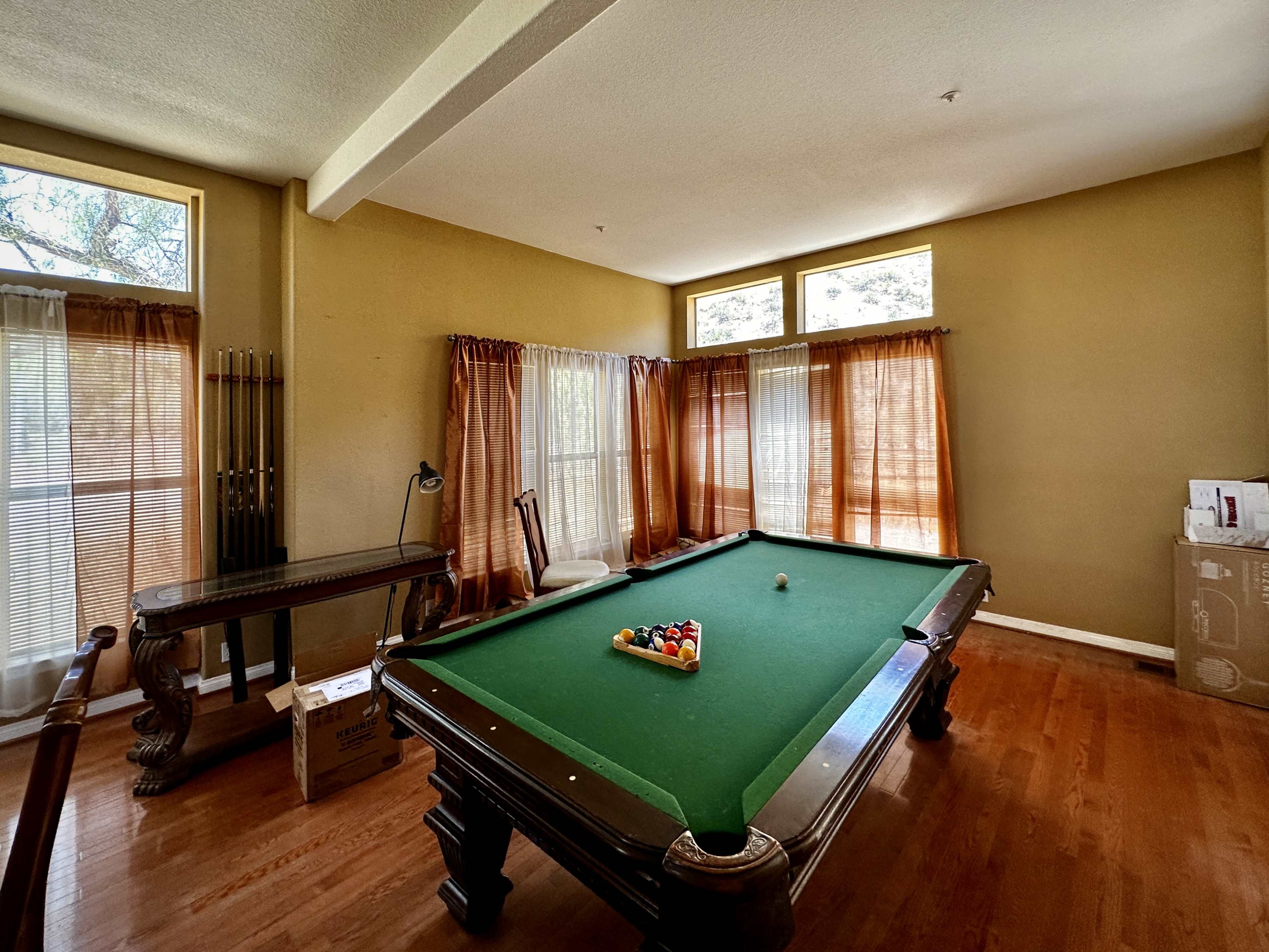 A room with a pool table, wooden flooring, and large windows with sheer curtains.