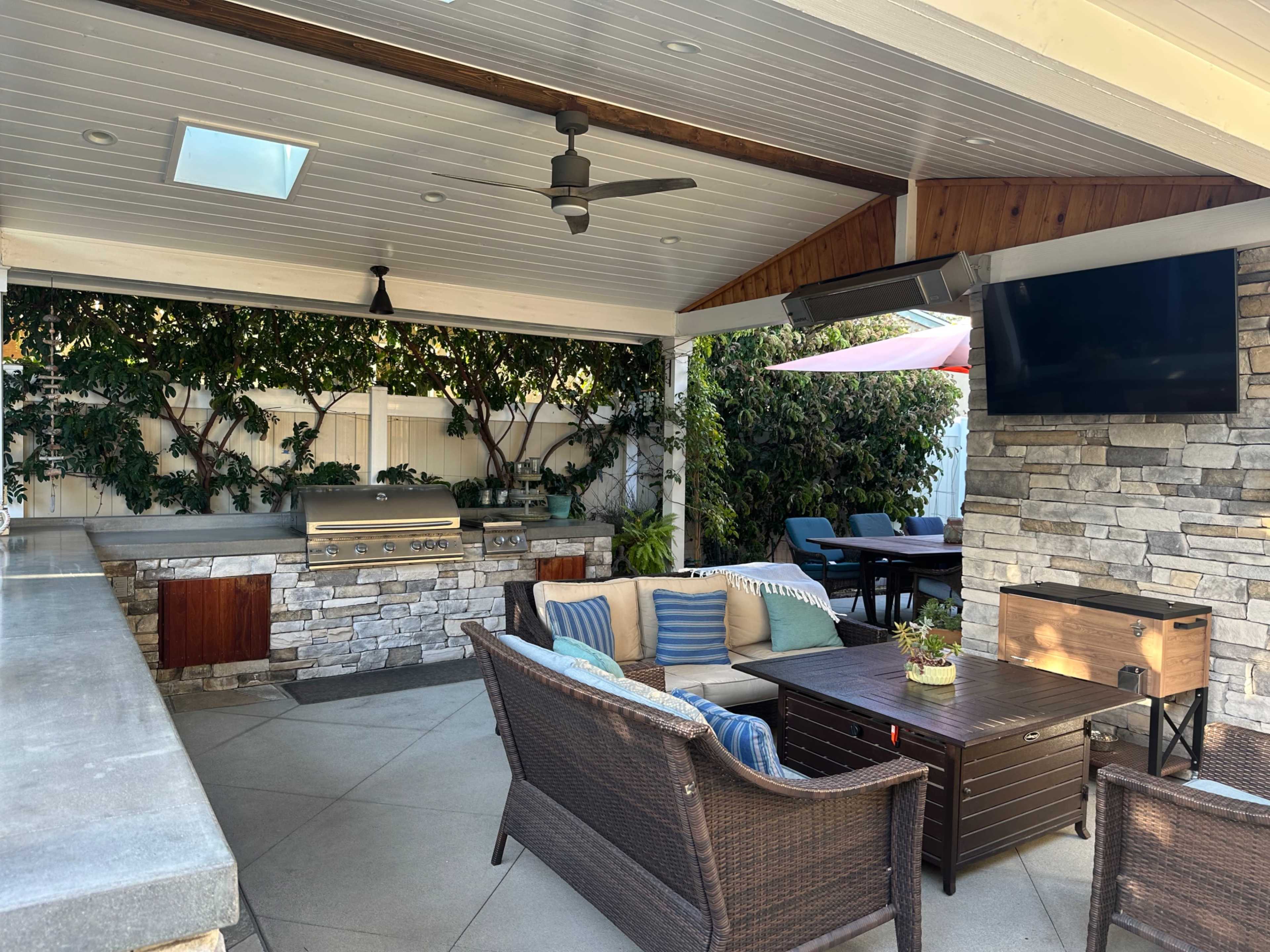 The image shows a covered outdoor patio area featuring a stone barbecue grill, seating with blue cushions, and a flat-screen television on the wall.