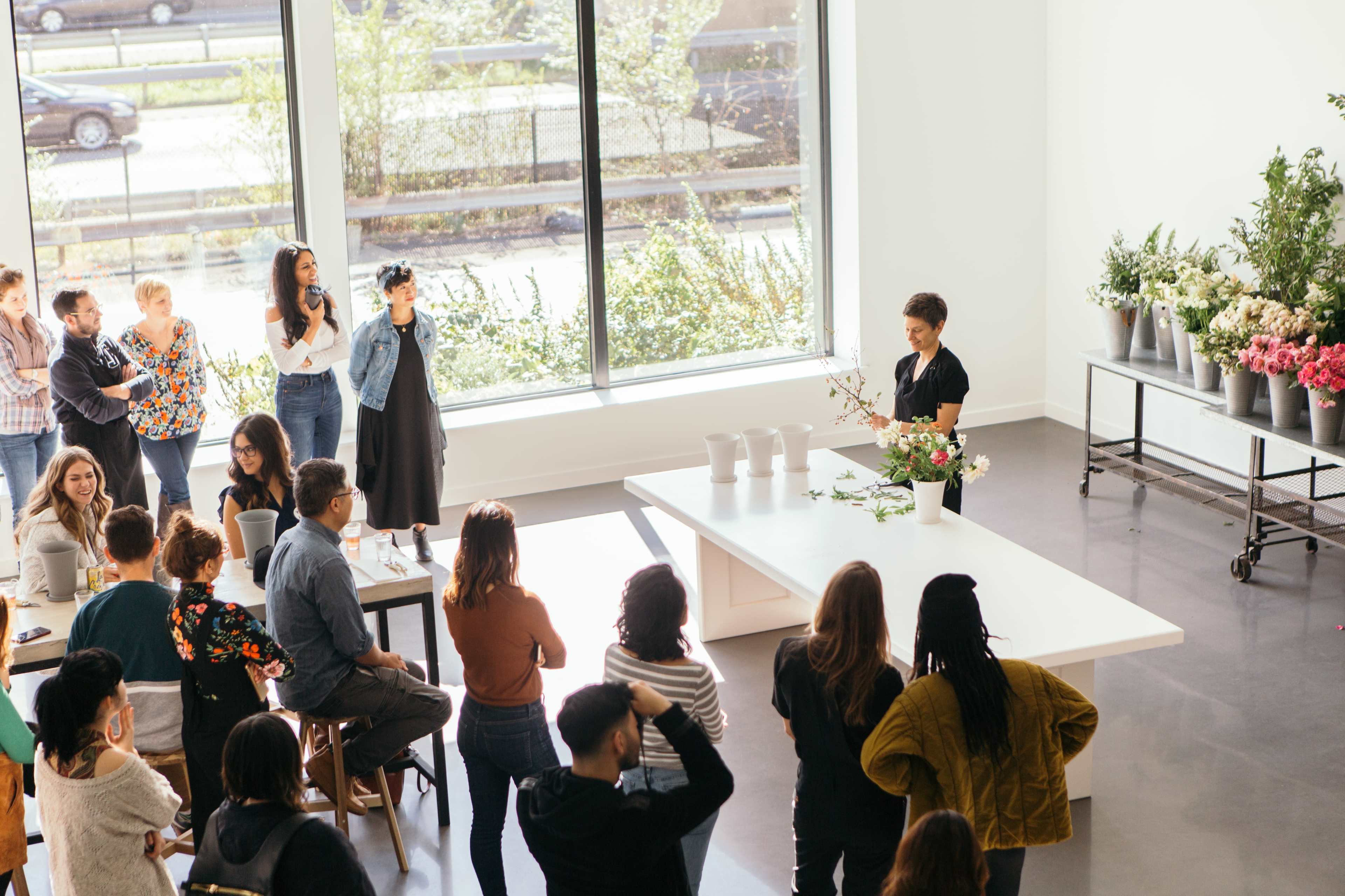 A group of people observes a floral arranging demonstration in a bright, spacious room with large windows.