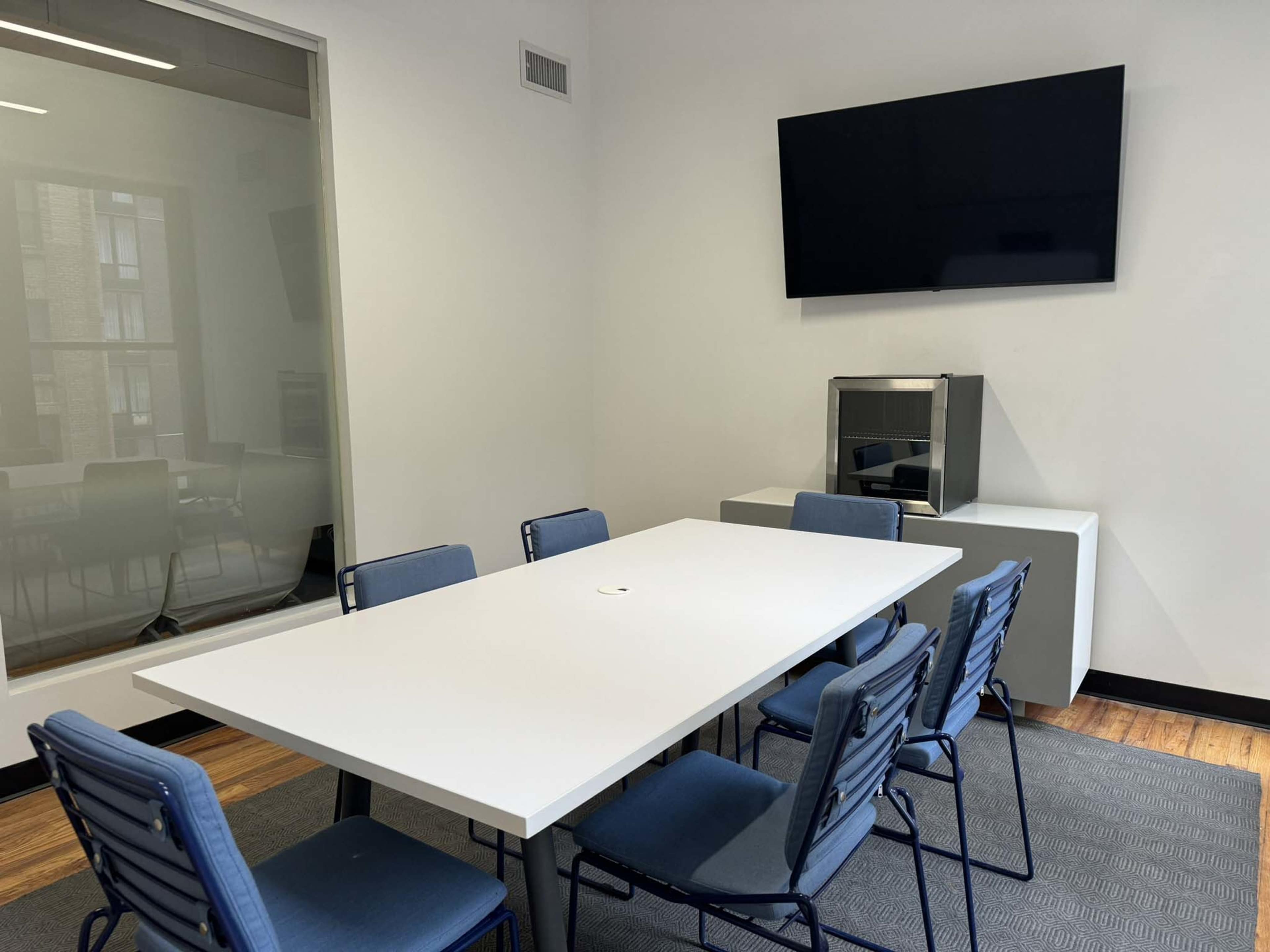A conference room with a long white table, blue chairs, a mounted television, and a small cabinet in a minimalist setting.
