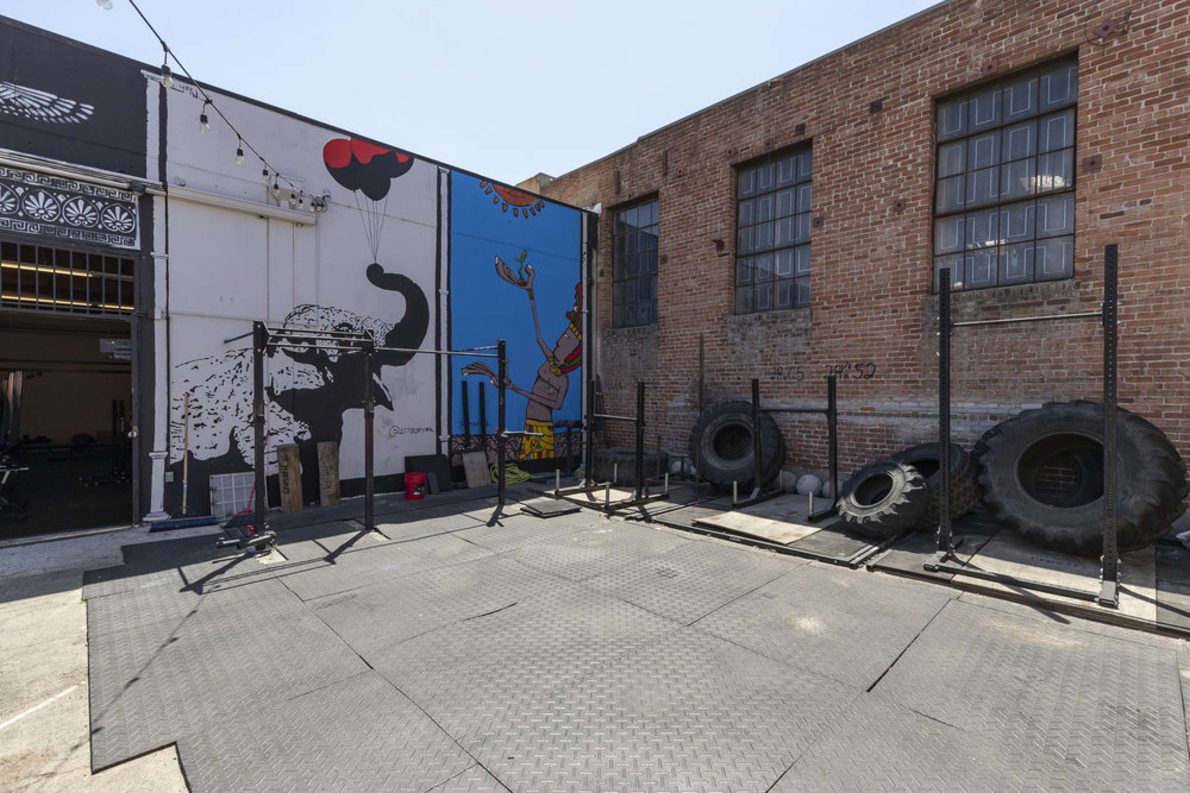 The image shows an outdoor workout area featuring gym equipment, large tires, and a colorful mural on a brick wall.