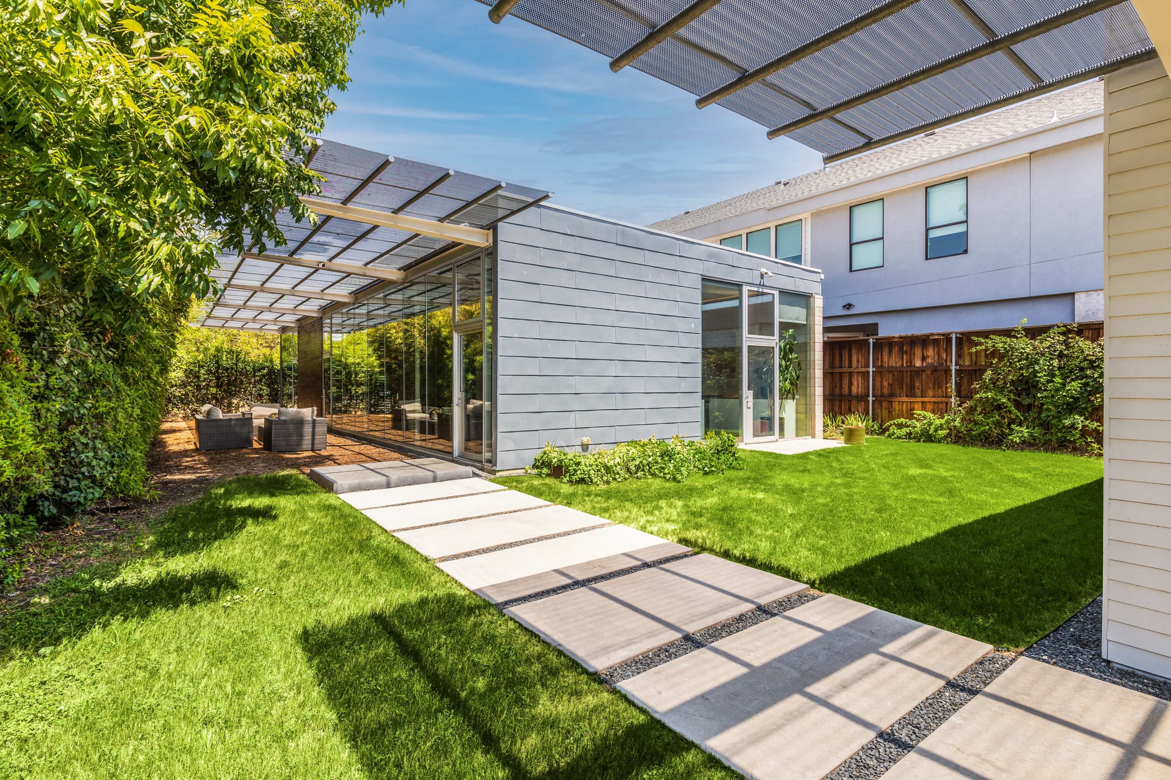 A modern garden space features a glass-walled building with a covered patio and a manicured lawn, surrounded by greenery.