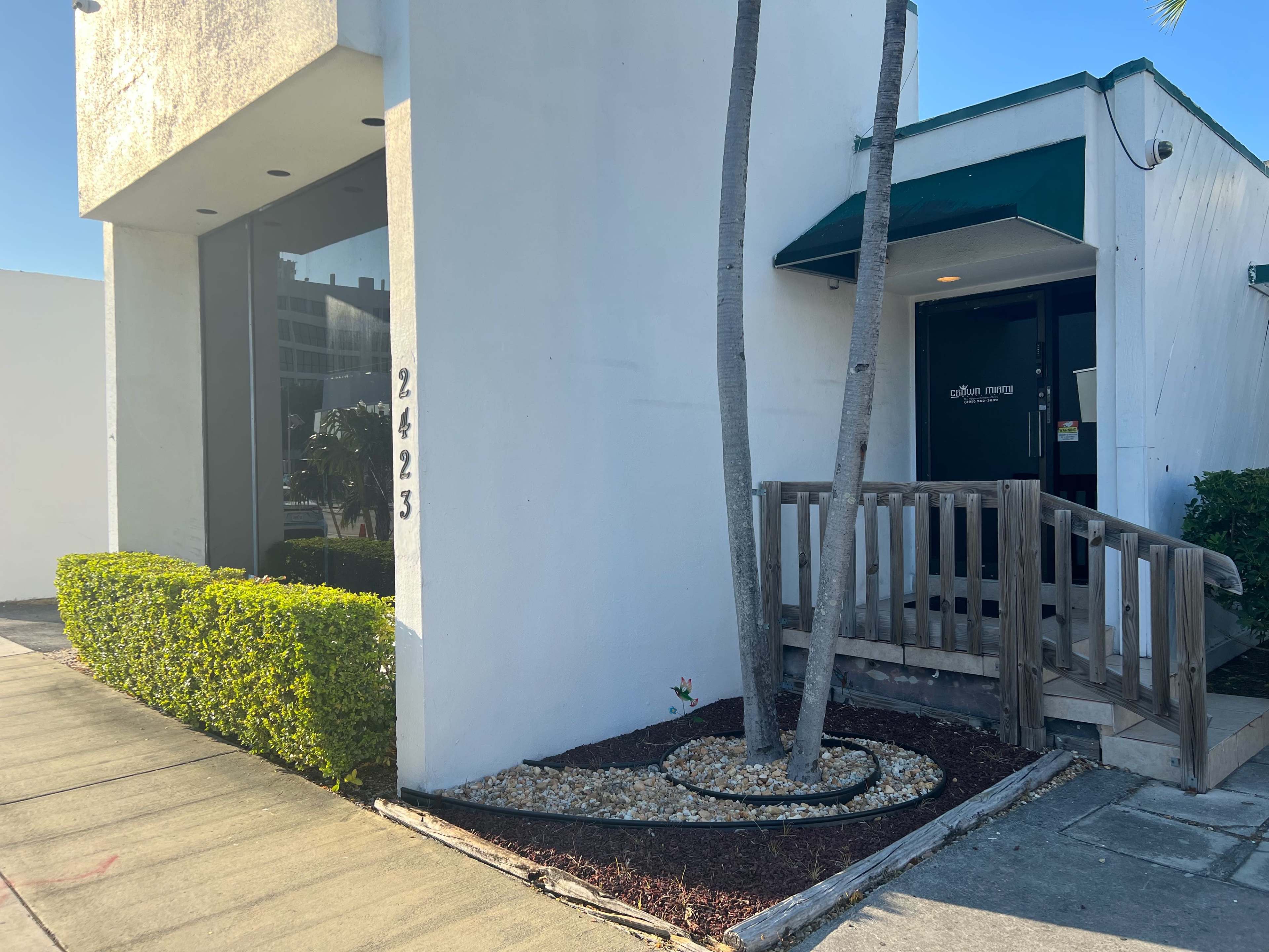 The image shows a corner building with the number 2423 displayed, featuring a ramp leading to a door and landscaped greenery nearby.