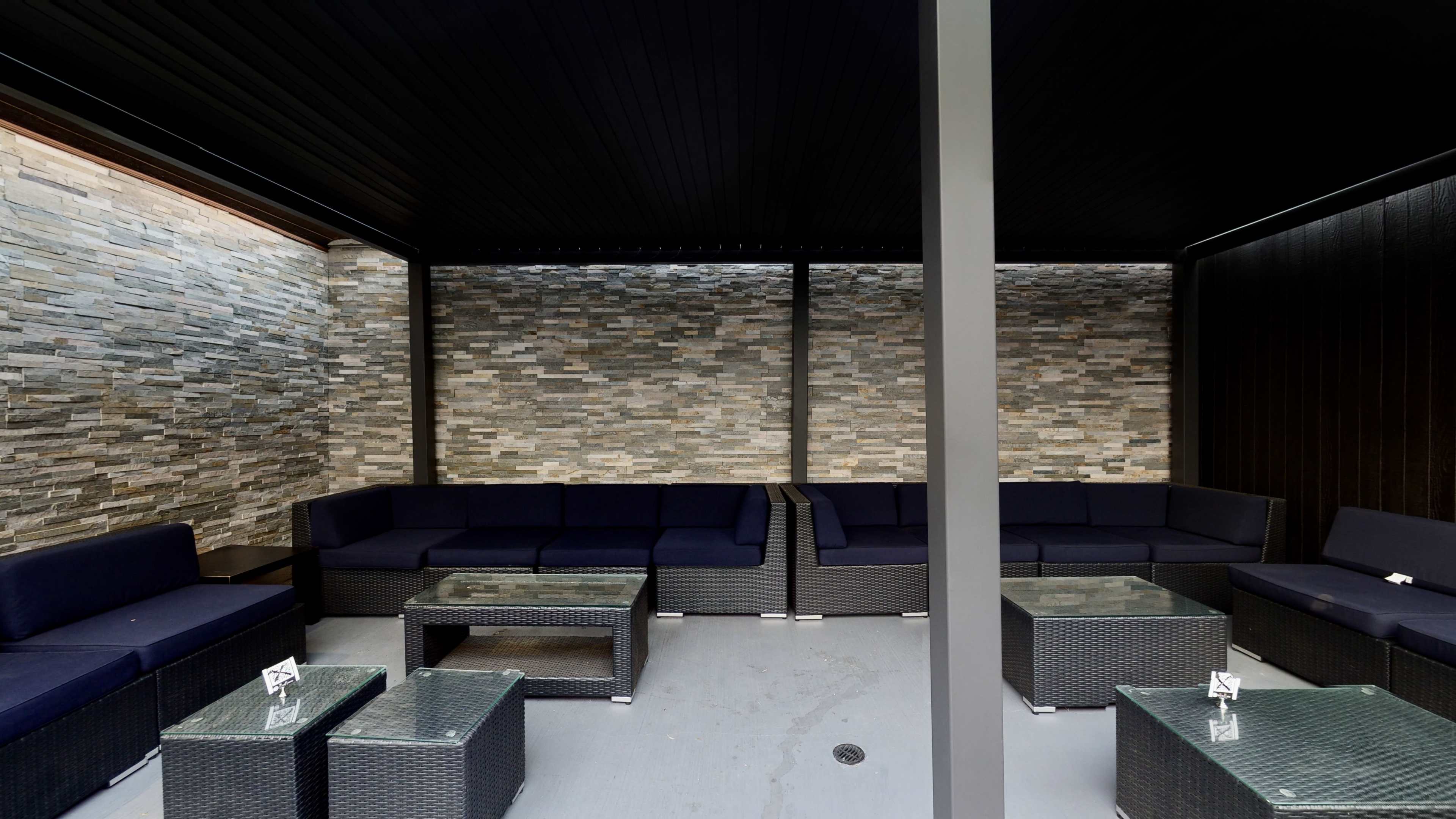The image shows a modern outdoor lounge area with dark wicker furniture, glass coffee tables, and stone-wall accents.