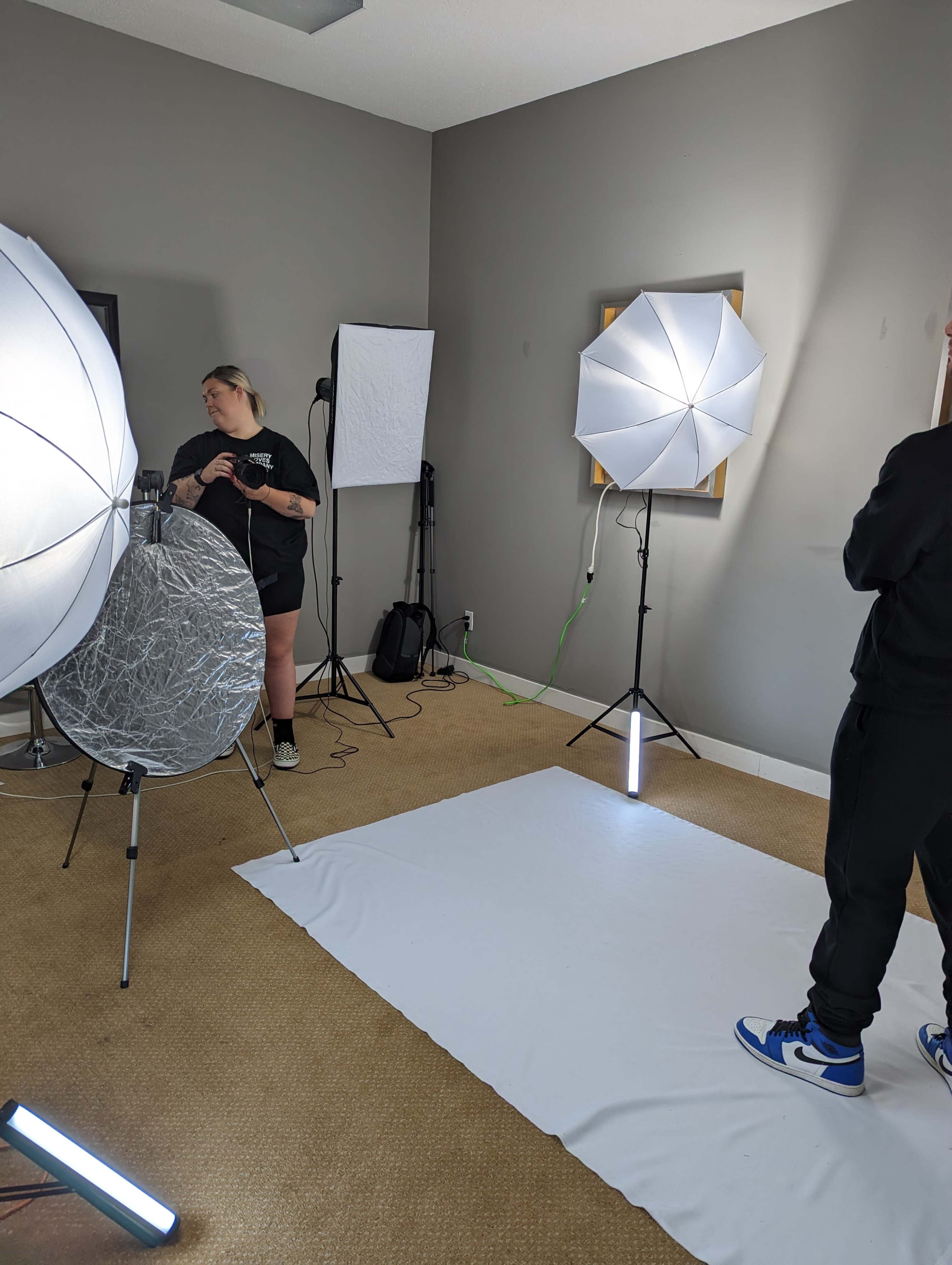 A photography studio is set up with softbox lights, a backdrop, and a model adjusting equipment.