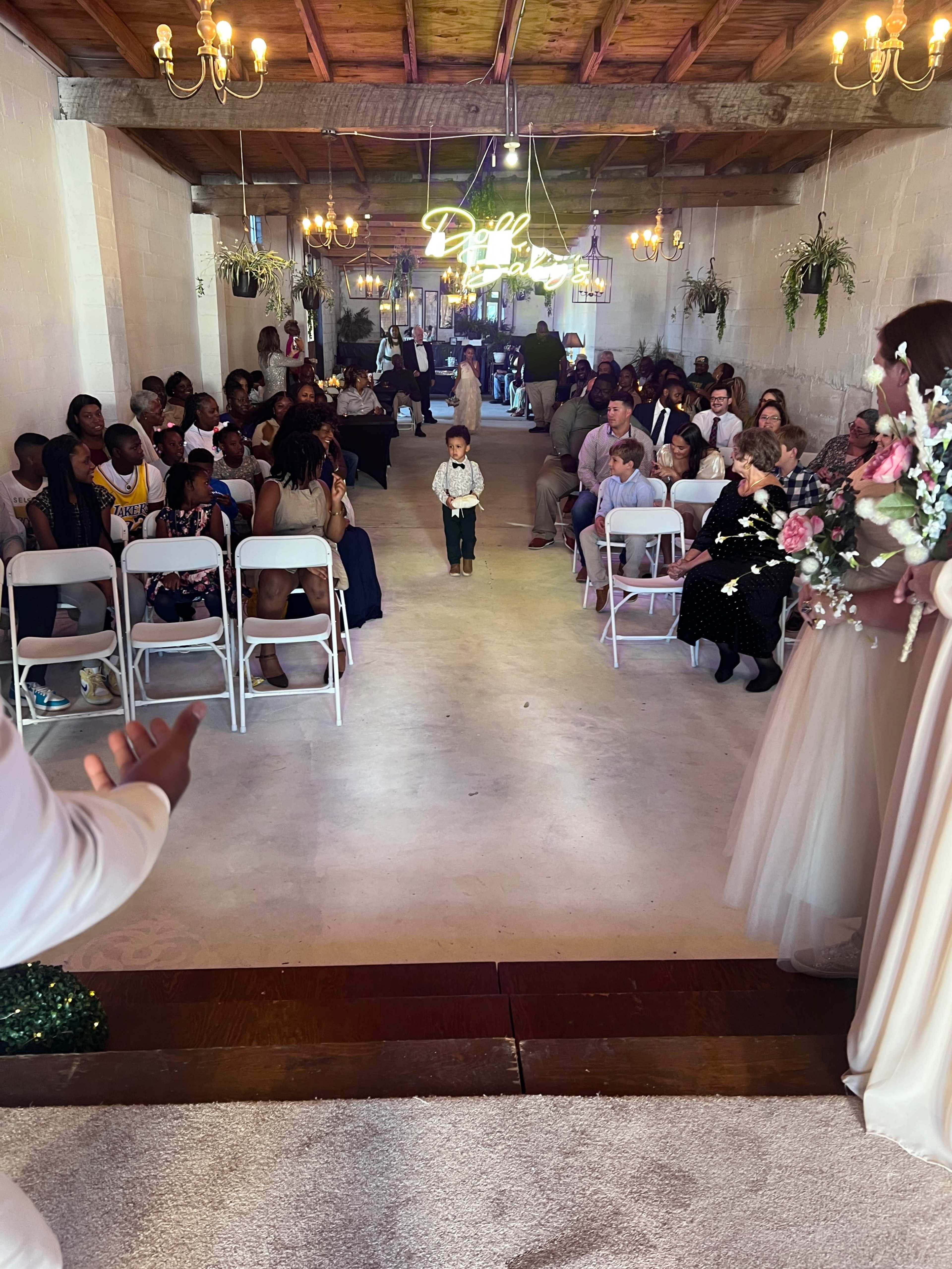 A child in formal attire walks down the aisle of a decorated venue filled with seated guests.