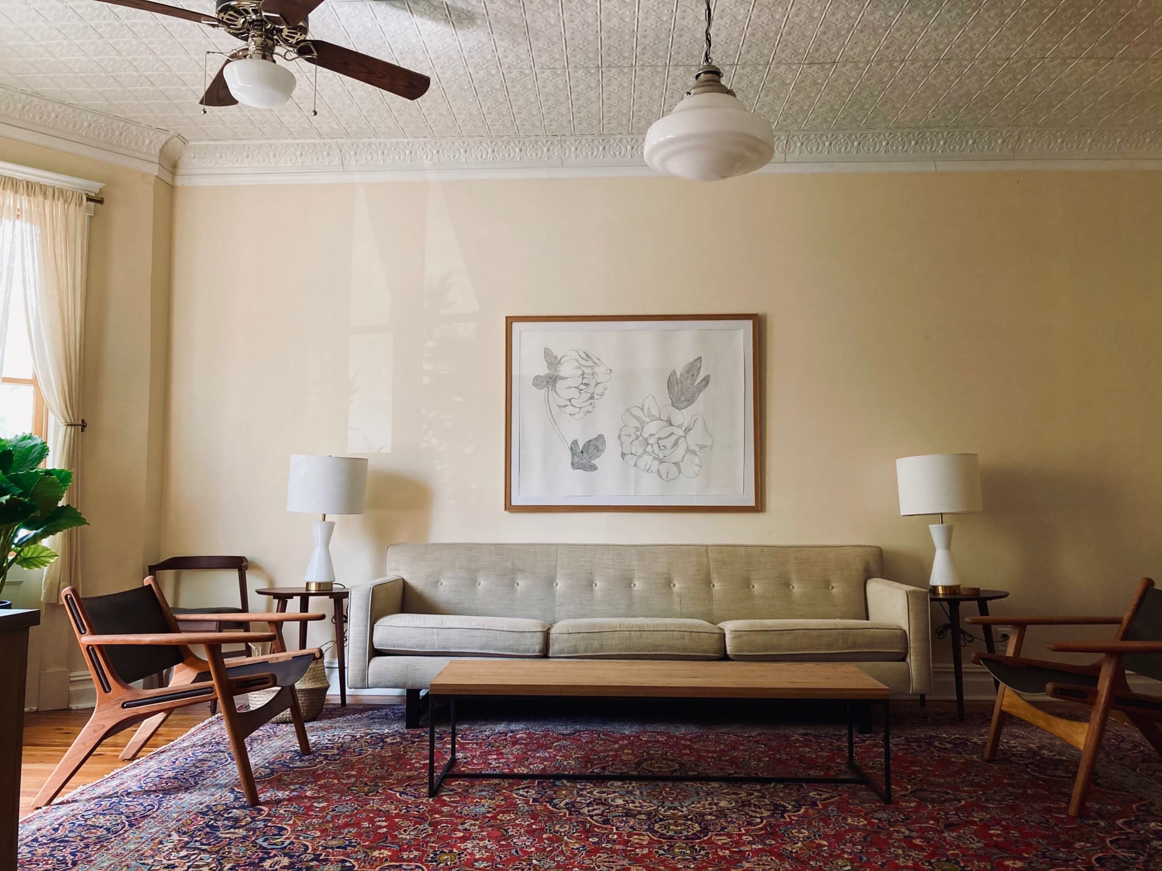 The image shows a living room with a sofa, two armchairs, a coffee table, and a framed artwork on the wall, accompanied by two floor lamps and a decorative plant.