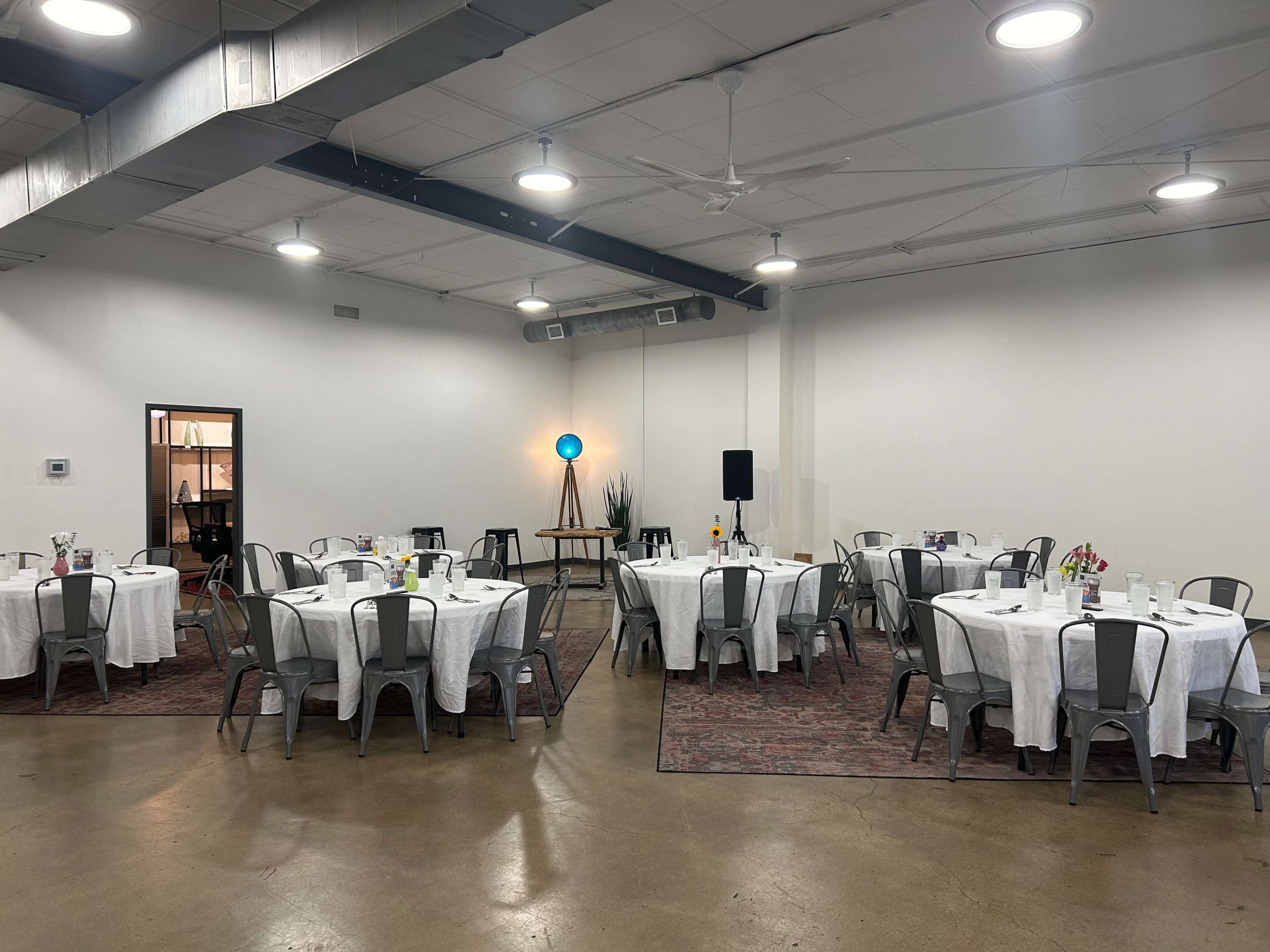A spacious event room features several round tables with white tablecloths and metal chairs, adorned with centerpieces, in a well-lit area.