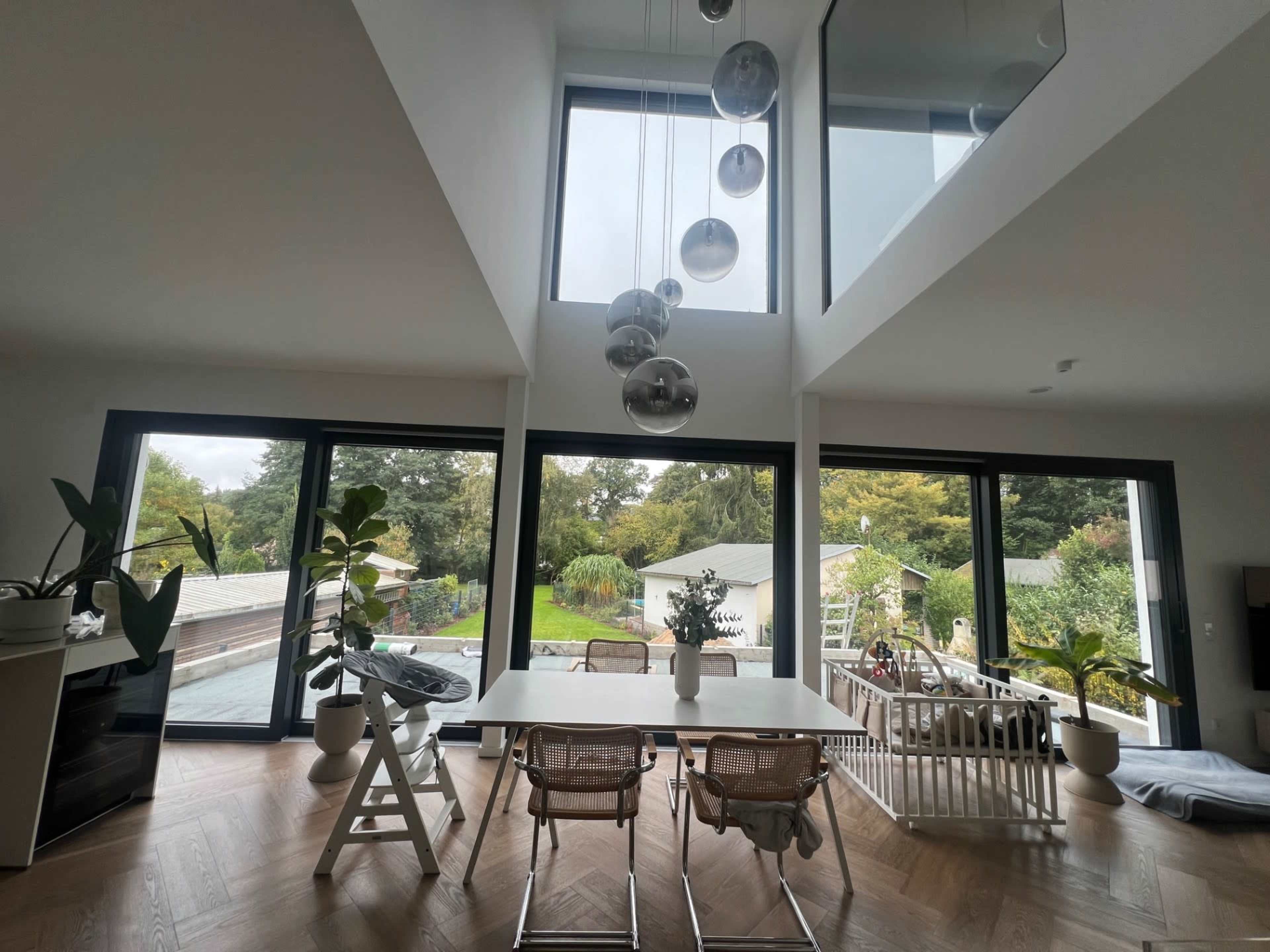The image shows a modern, open-concept living space with large windows, a dining table, and greenery, overlooking a backyard.