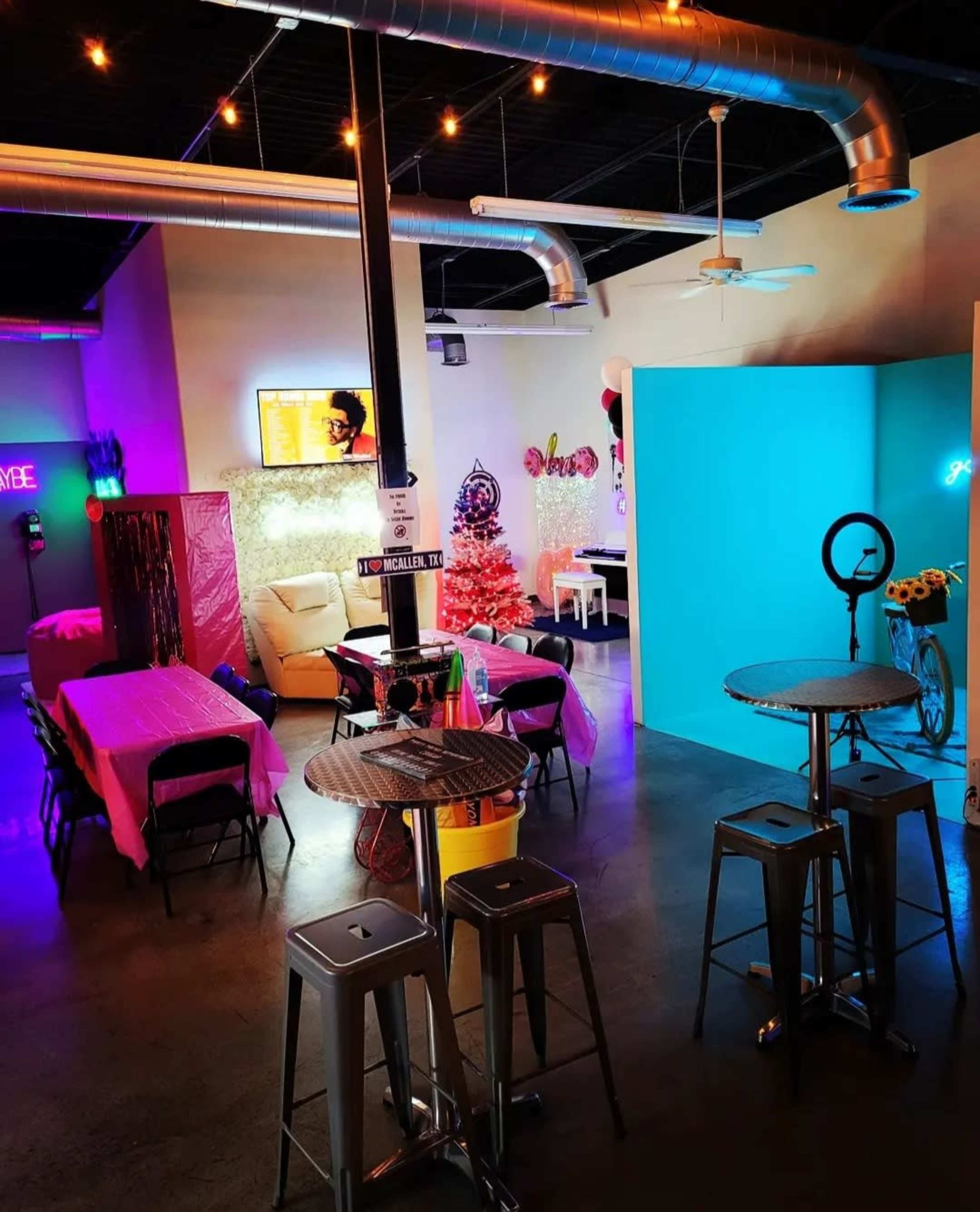 A bright event space featuring tables with pink tablecloths, a decorated Christmas tree, and a vibrant blue wall, with bar stools and various decorations.