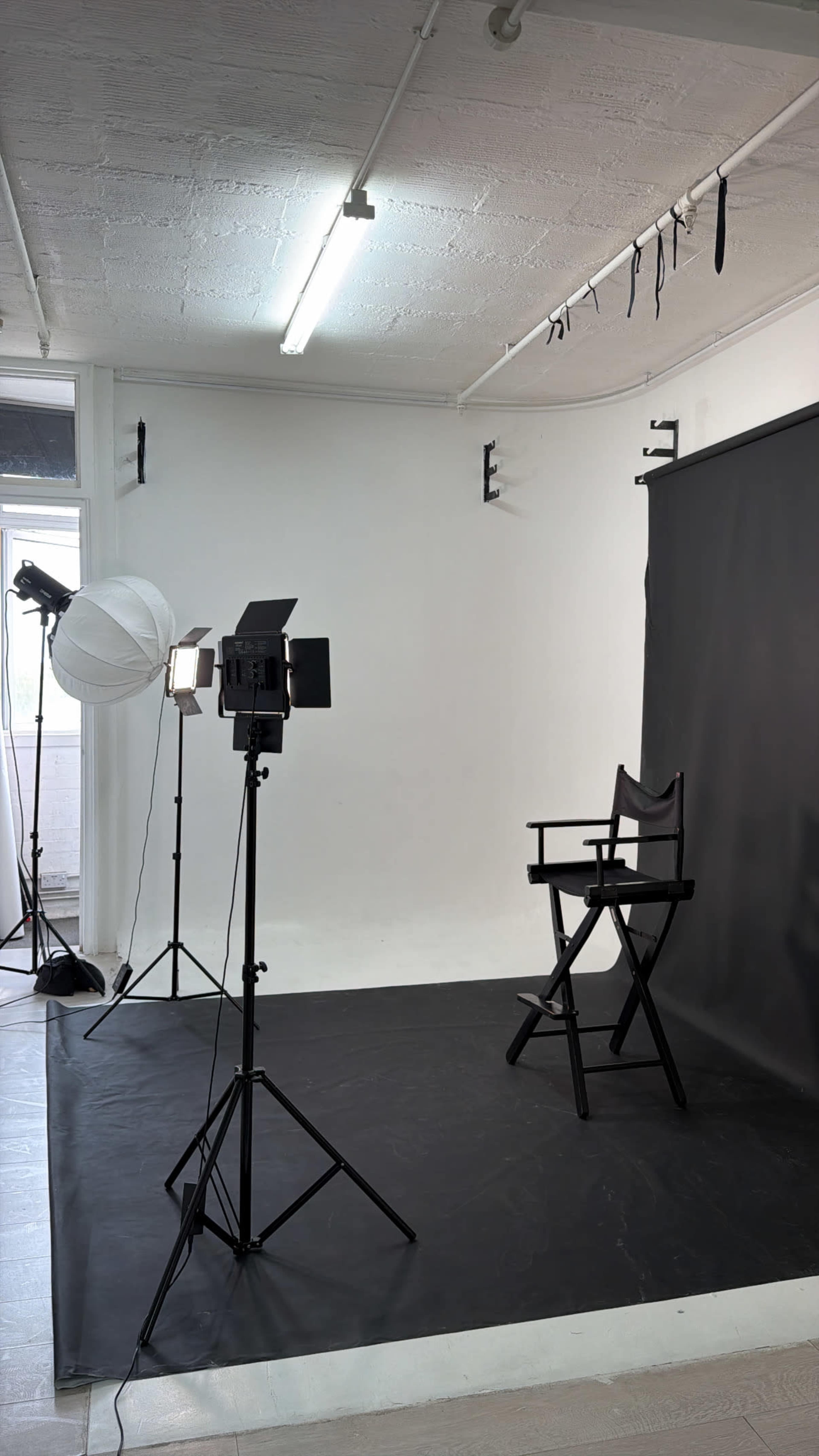 The image shows a photography studio with a black backdrop, a director's chair, and multiple lighting equipment set up for a photoshoot.