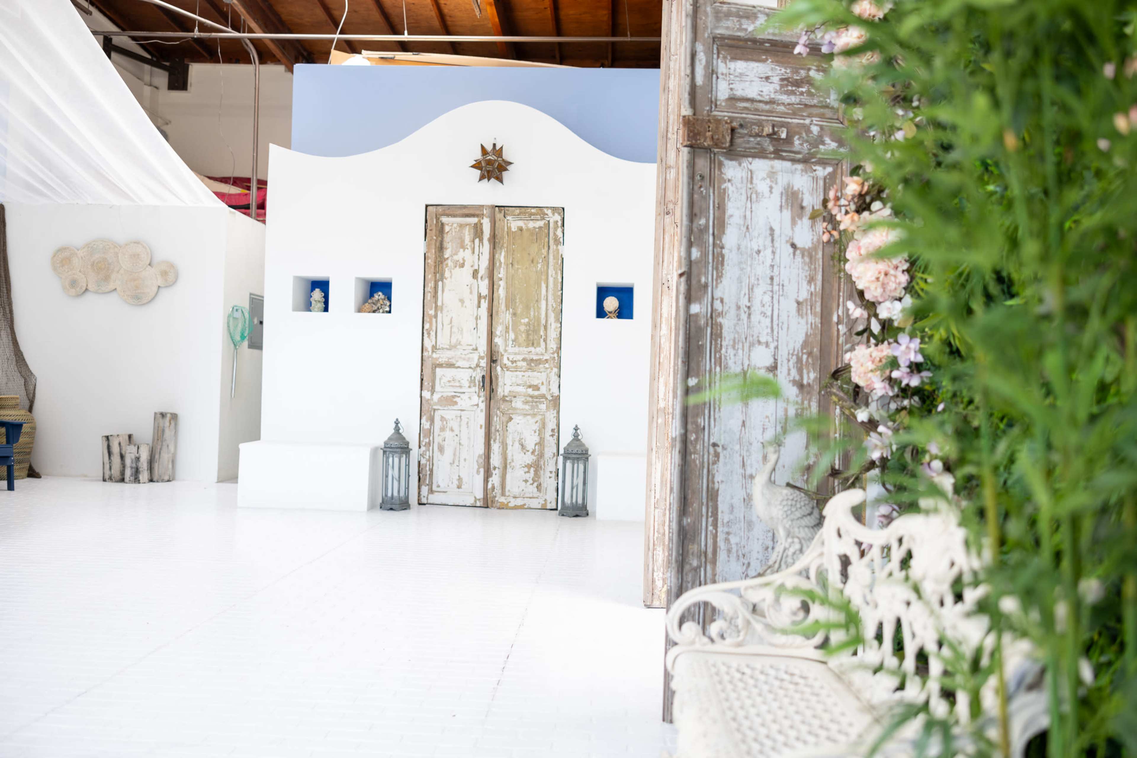The image depicts a bright, airy interior with a weathered wooden door, blue wall accents, and decorative elements, surrounded by greenery and soft lighting.
