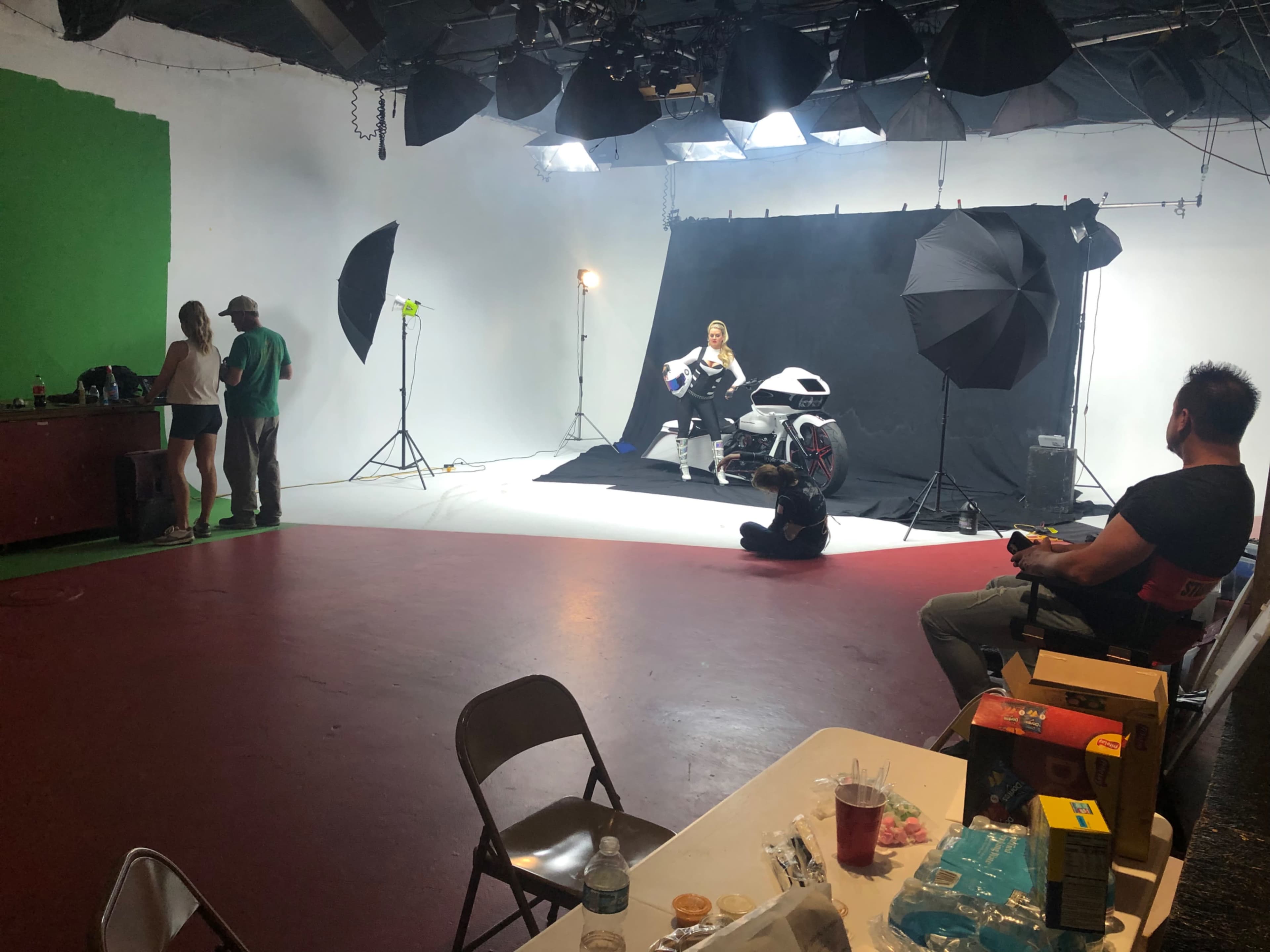 A photoshoot is underway in a studio with a model posing on a motorcycle, while crew members work in the background.