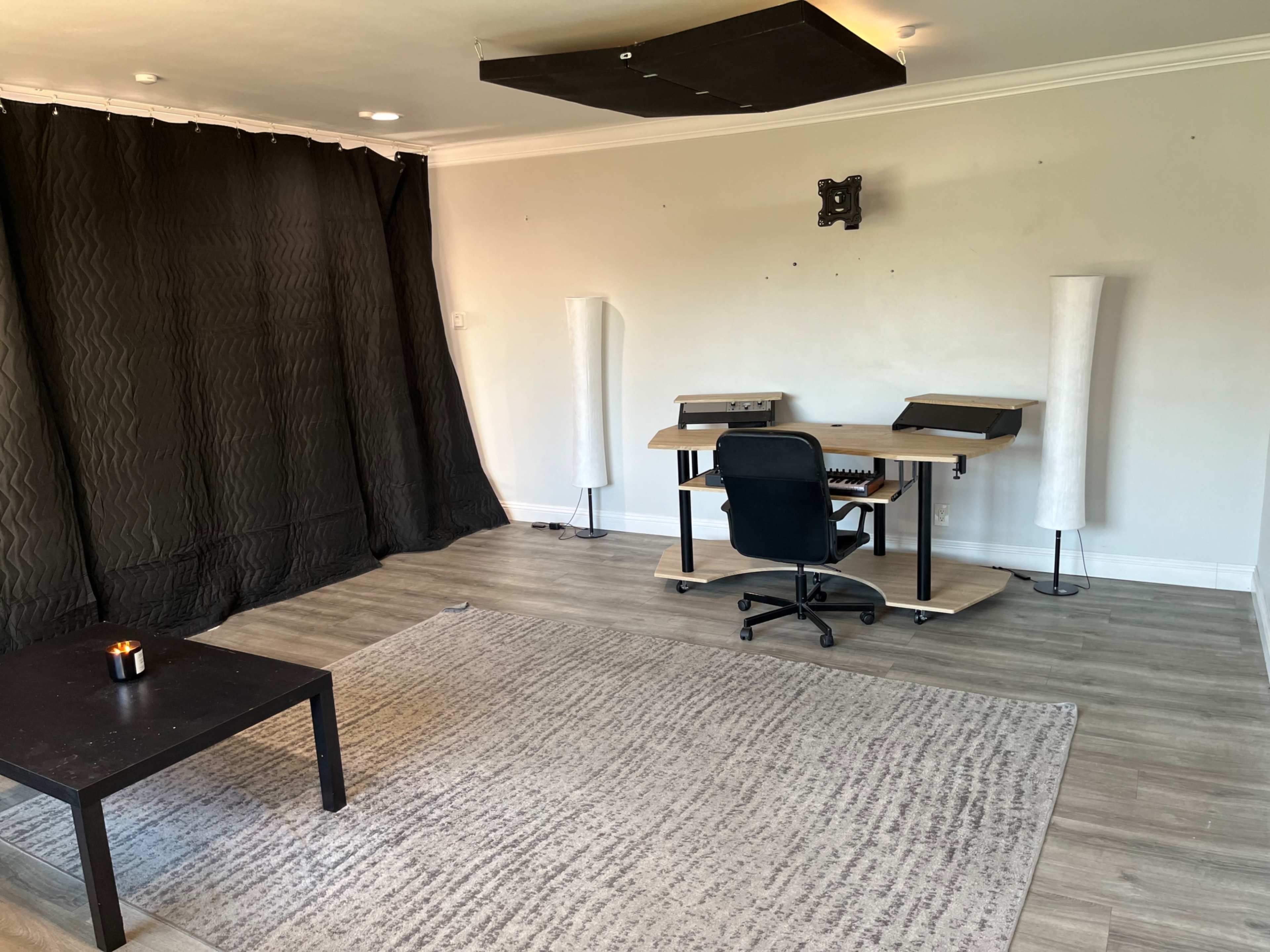 The image shows a partially furnished room featuring a black curtain, a desk with a keyboard, a chair, and two floor lamps, along with a small table and a rug on the floor.