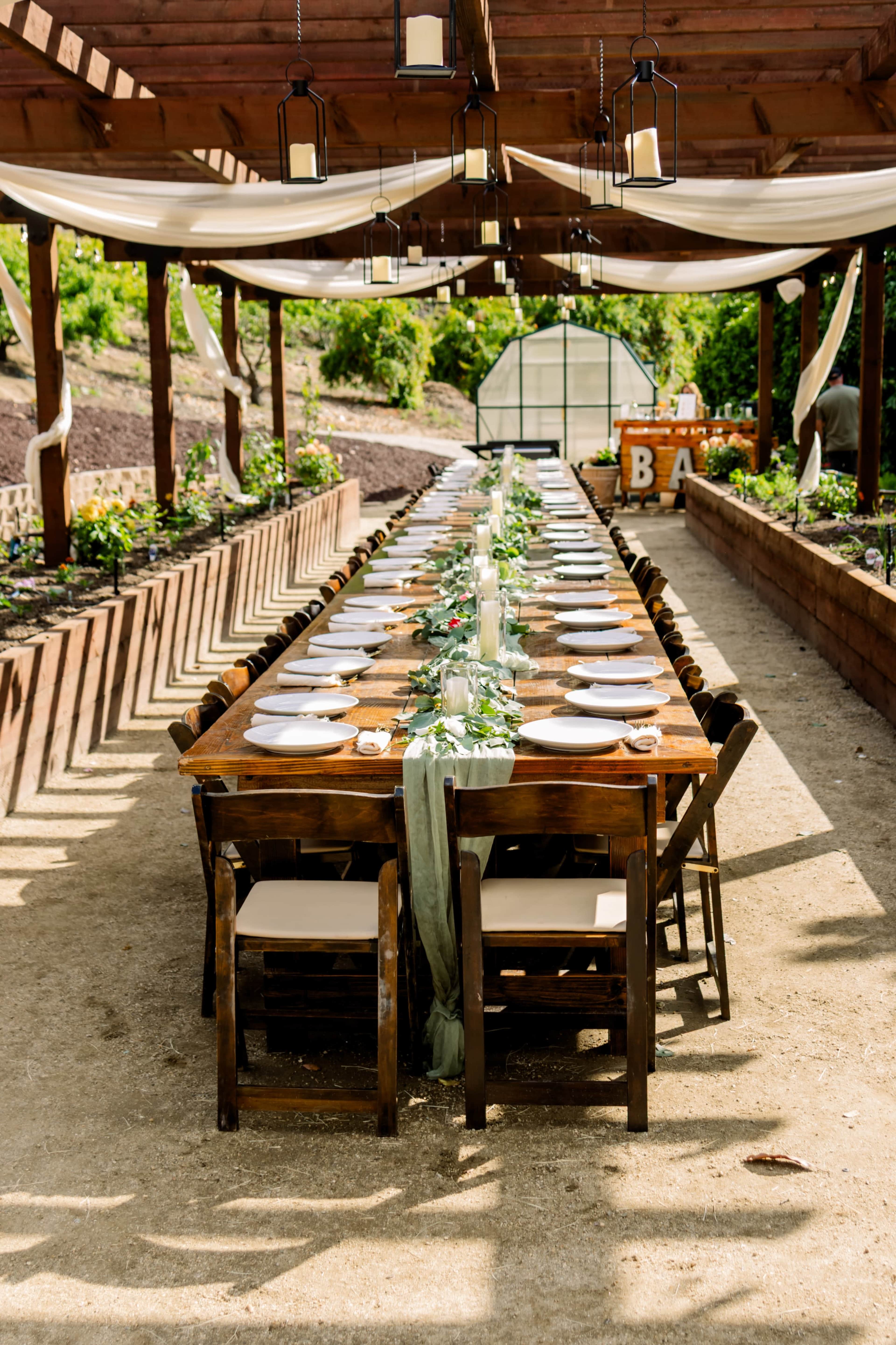 A long wooden table is set for a meal outdoors, lined with plates and greenery decorations under a canopy of draped fabric and hanging lights.