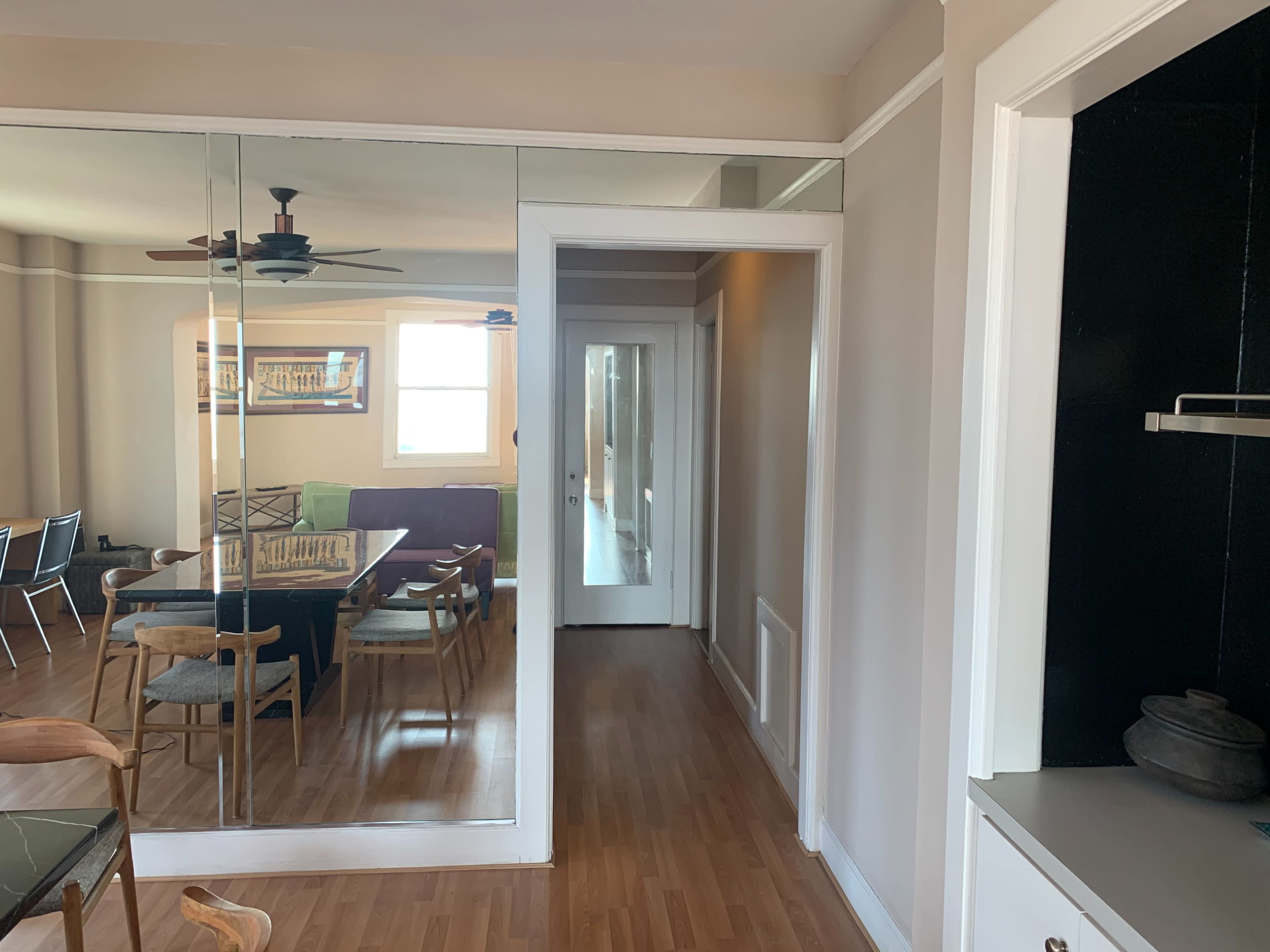 A hallway leading to a door, with a mirrored wall on one side and a dining area visible in the background.